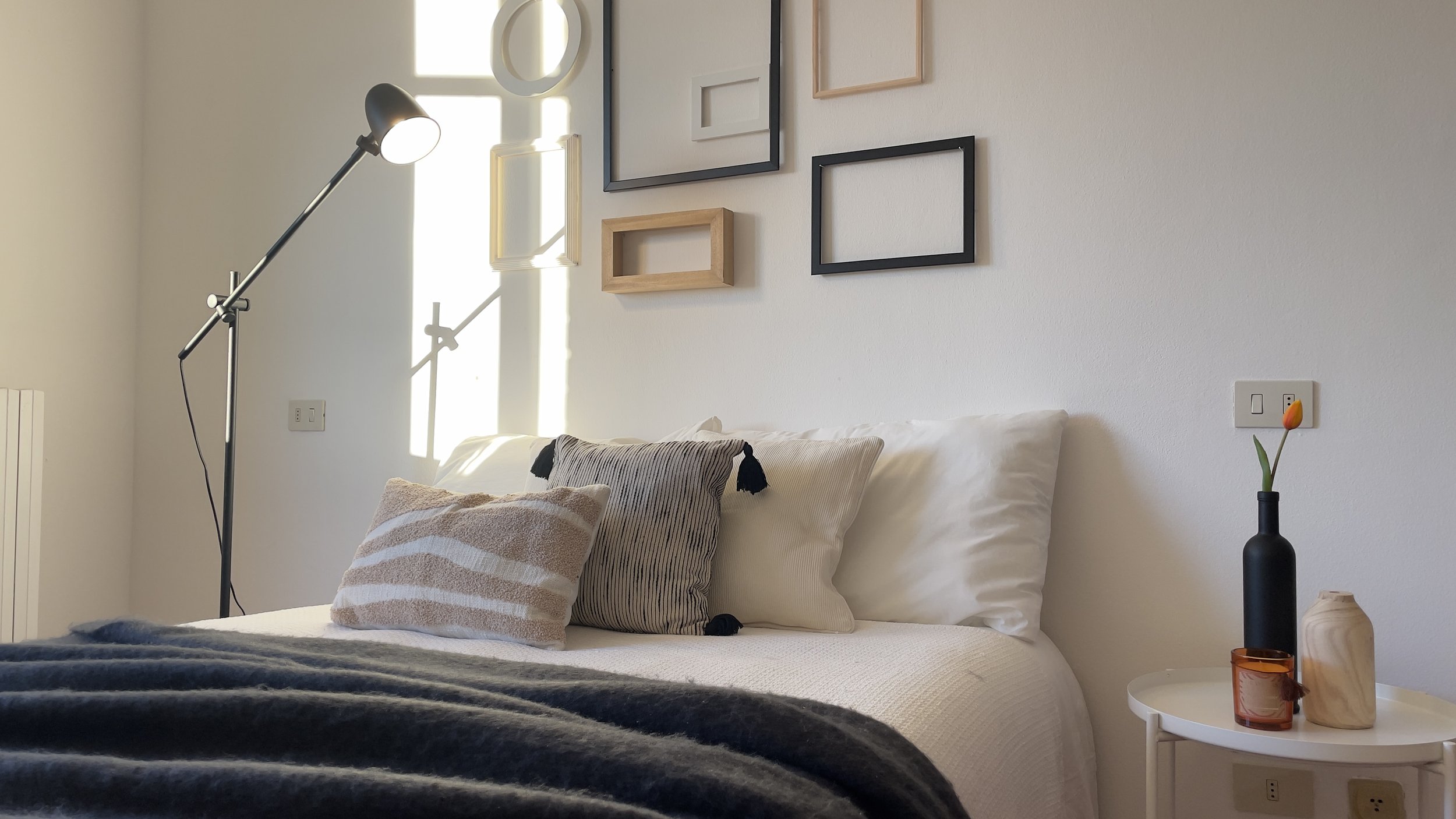 Camera da letto moderna con letto ben fatto, cuscini decorativi, parete con cornici vuote, lampada da lettura nera, tavolino con vaso nero, candela e piccola caraffa di legno.