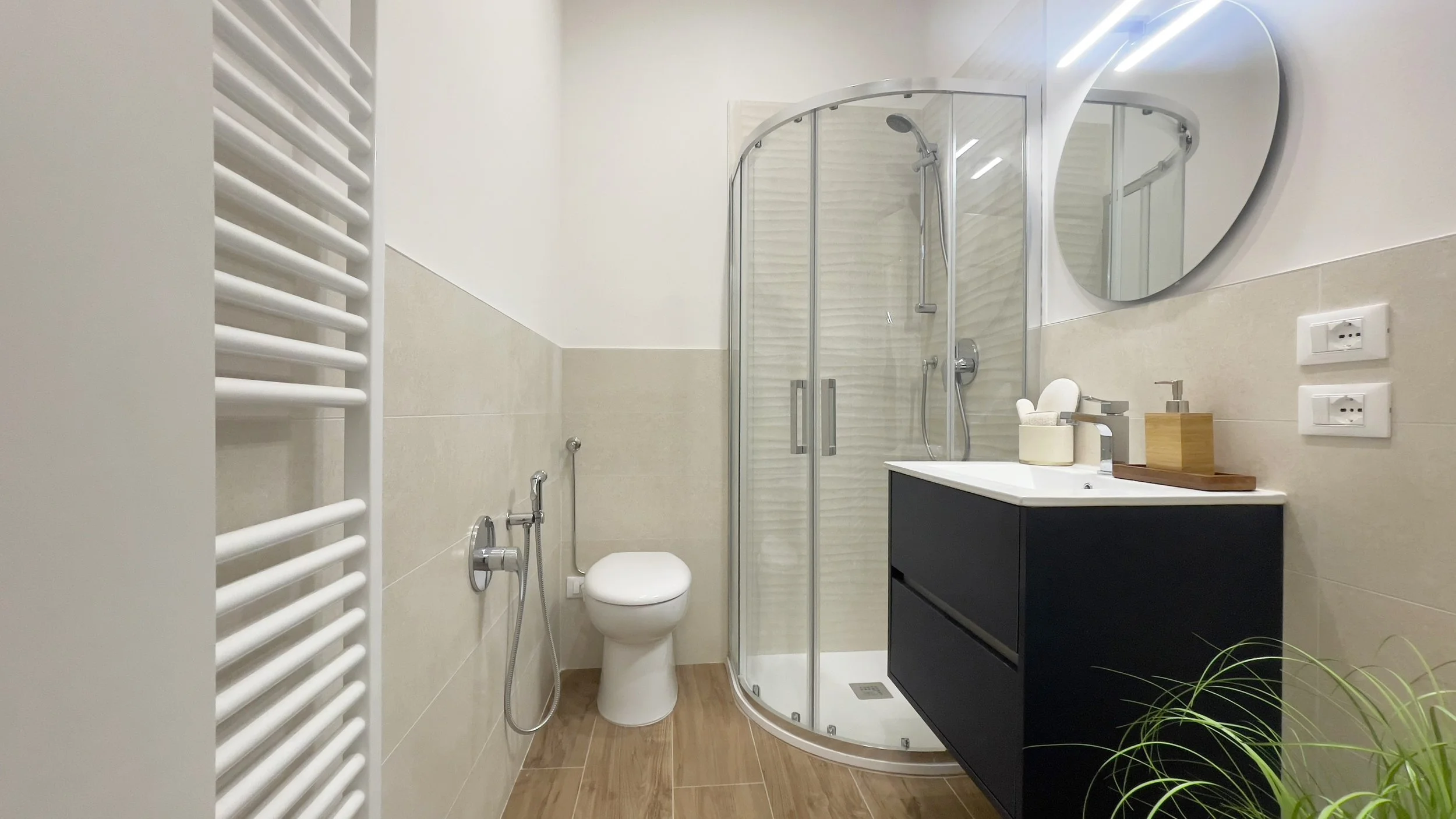 Bagno moderno con doccia in vetro, lavabo nero con specchio rotondo, wc, e scaldasalviette bianco.