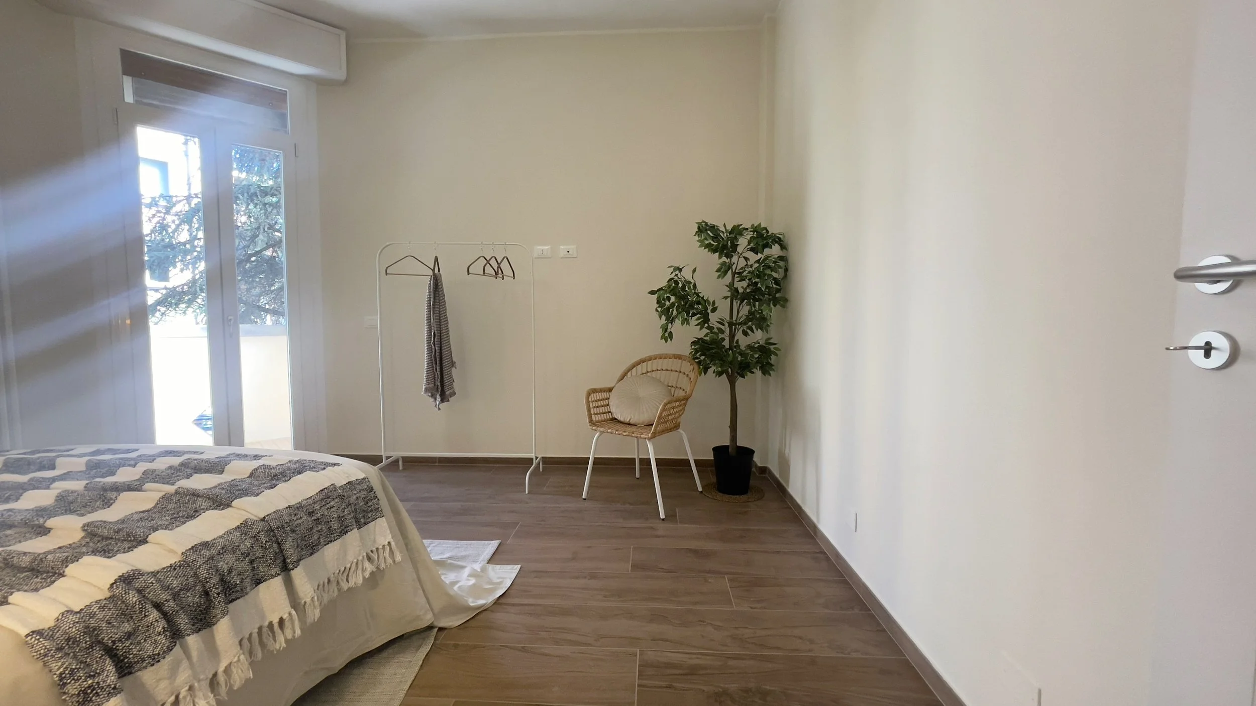 Camera minimalista con un letto, appendiabiti, sedia di vimini, pianta in vaso e una finestra che lascia entrare luce naturale.
