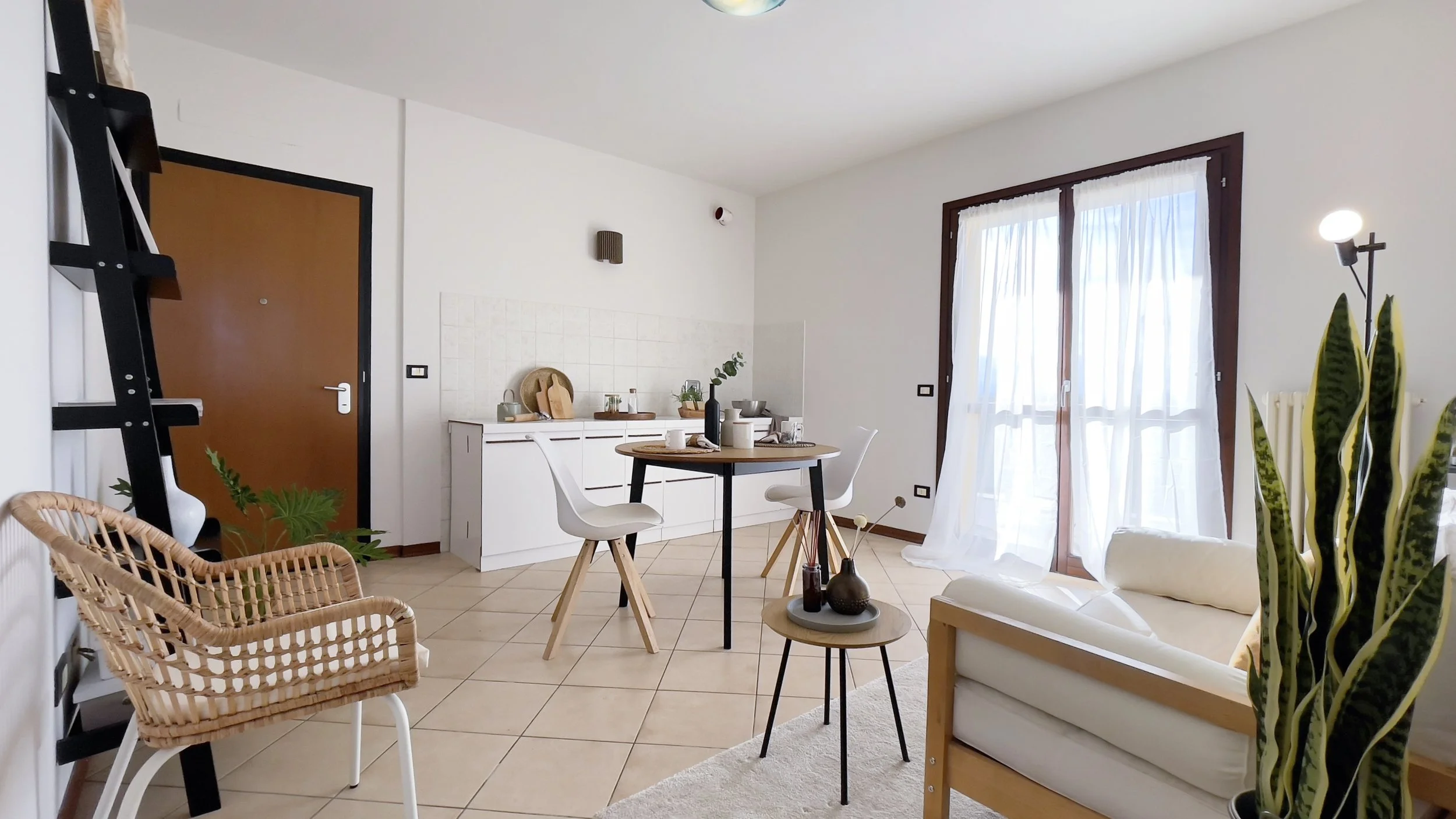Soggiorno moderno con tavolo rotondo e sedie bianche, cucina minimalista, porta d'ingresso in legno, poltrona in vimini, ripiani con decorazioni, pianta e lampada, finestra con tende bianche.