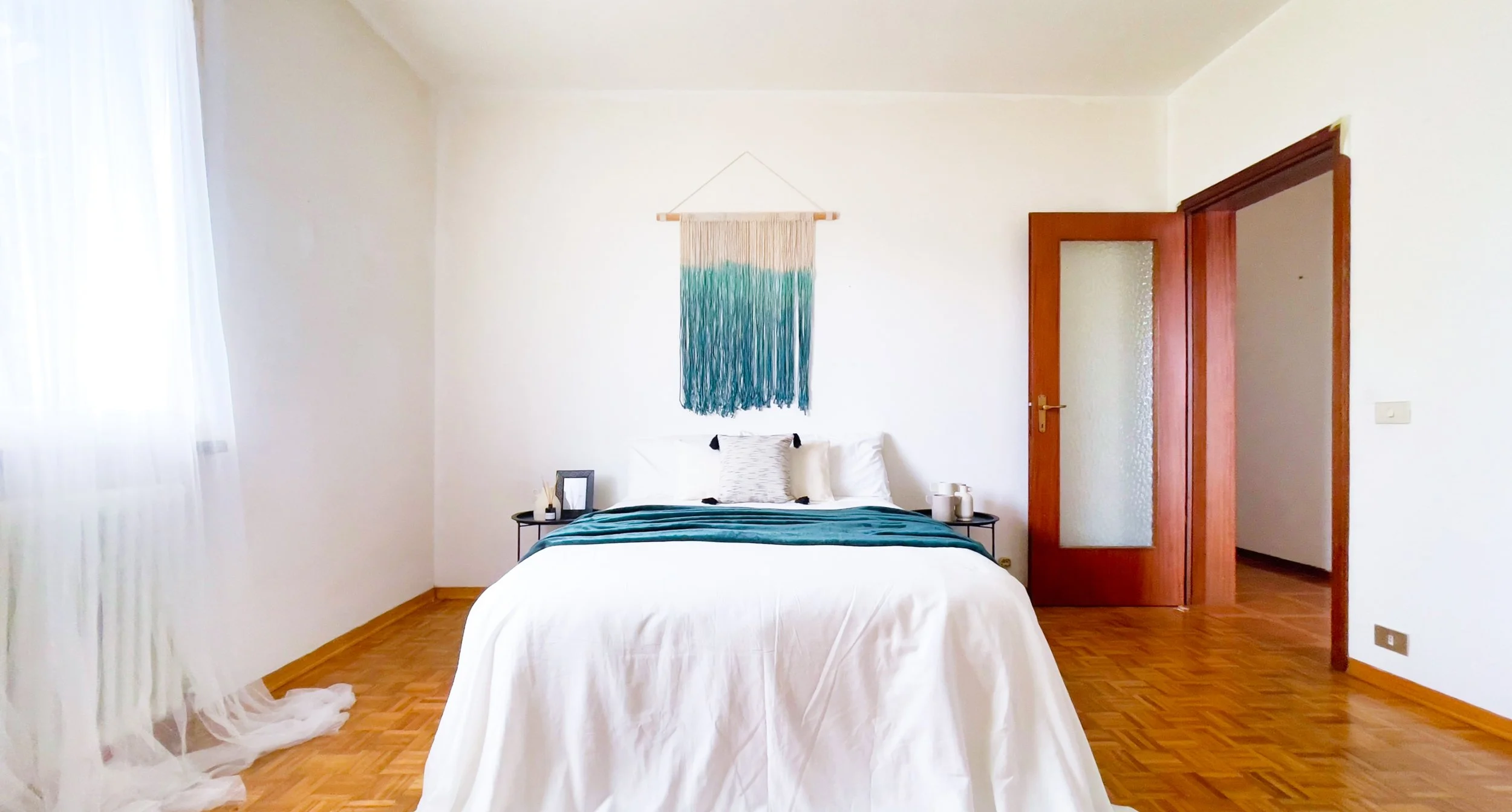 Camera da letto minimalista con letto matrimoniale, coperta turchese, arte tessile appesa al muro, pavimento in legno.
