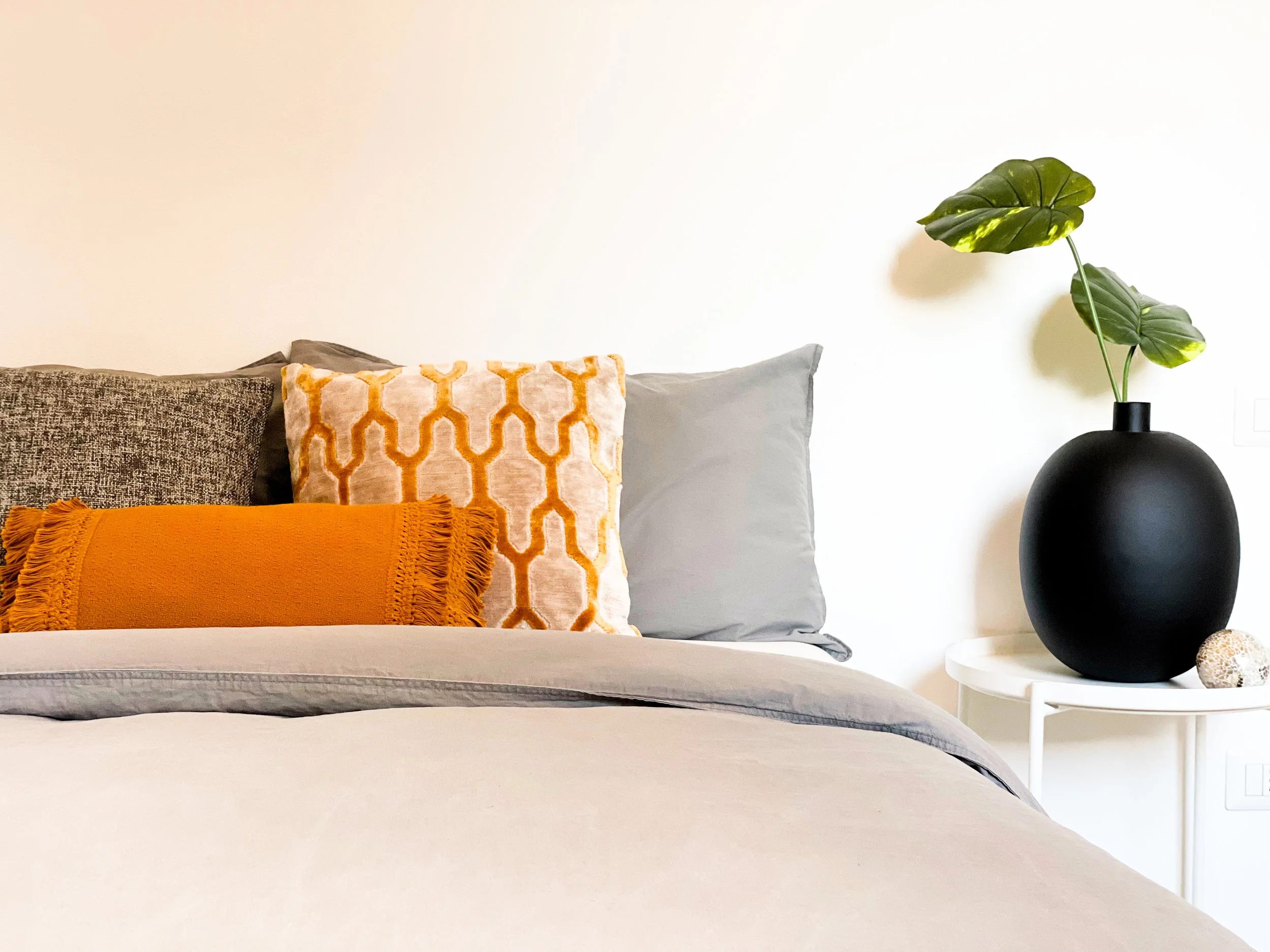 Camera da letto con letto e cuscini decorativi colorati vicino a un comodino con vaso nero e pianta.