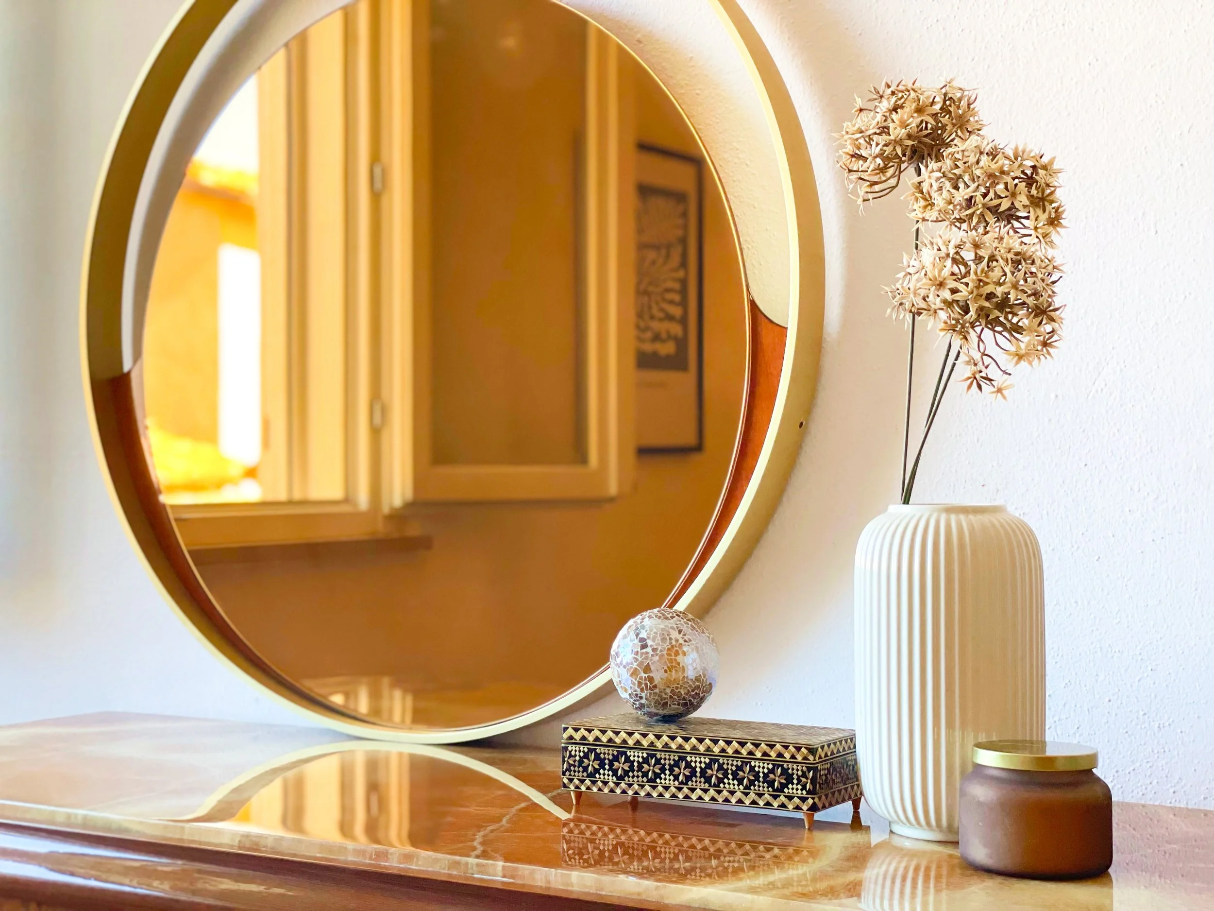 Composizione decorativa con specchio rotondo, vaso bianco con fiori secchi, scatola ornata e candela su superficie di legno.
