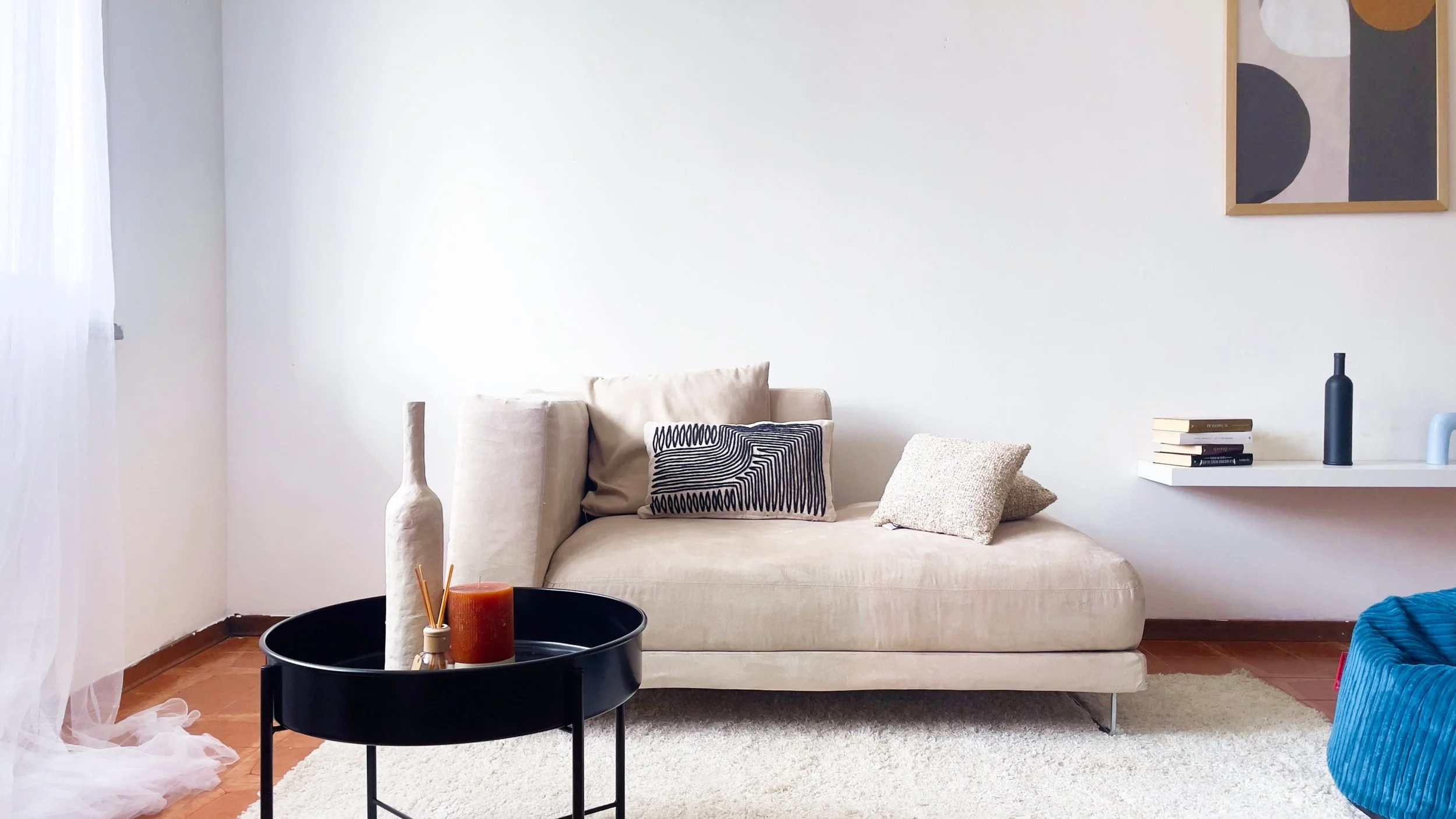 Soggiorno con divano beige, cuscini decorativi, tavolino nero con candele e deco, mensola con libri e oggetti decorativi, pouf blu, quadro astratto sul muro.