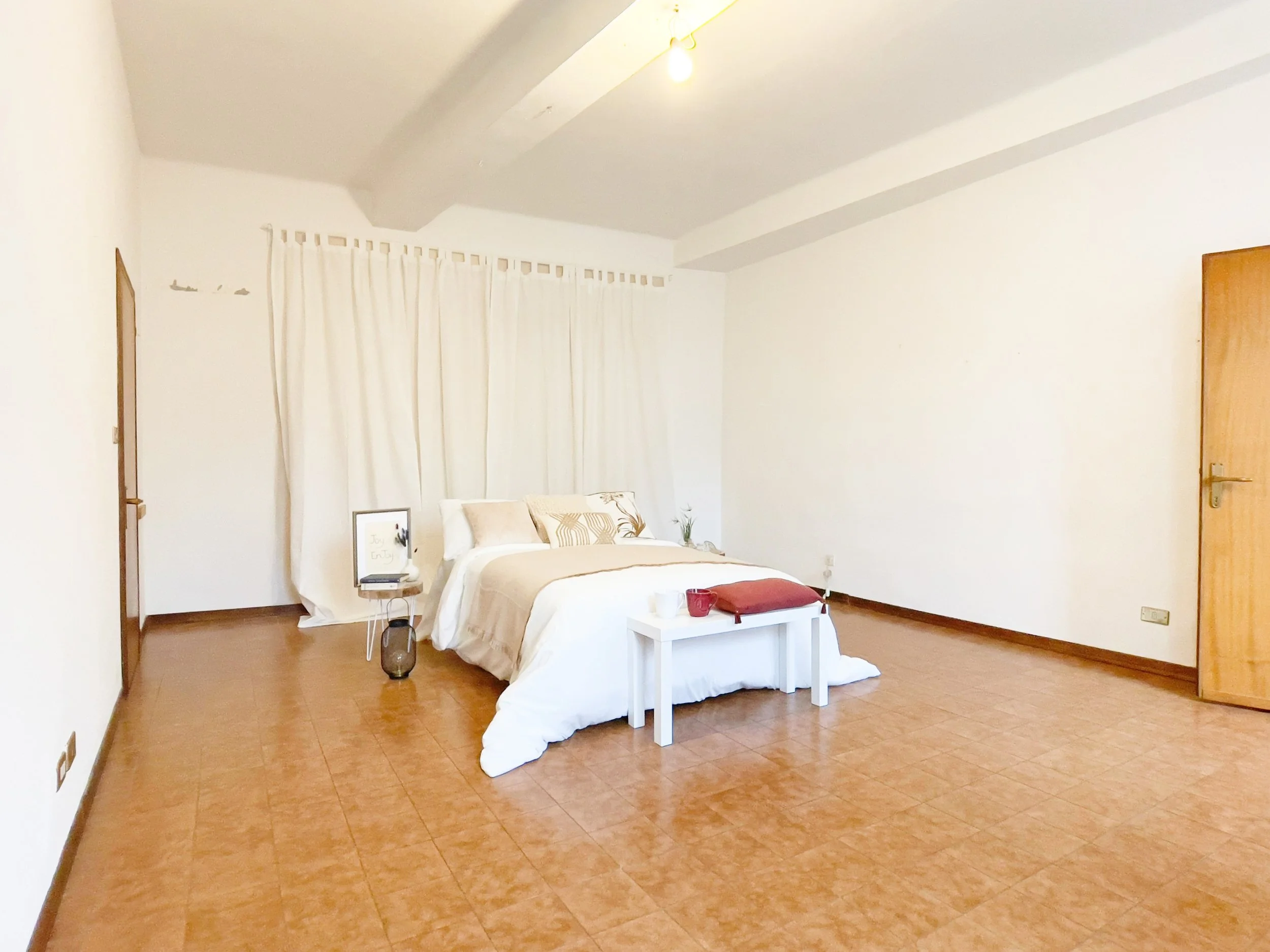 Camera da letto minimalista con letto matrimoniale, coperta bianca, tavolino su cui appoggiano oggetti decorativi e tendaggi bianchi sullo sfondo.
