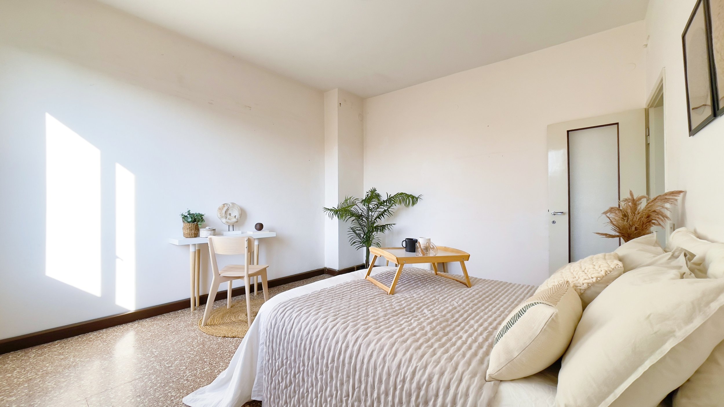 Camera da letto luminosa con letto, coperte, cuscini e un vassoio da colazione, scrivania con sedia e pianta decorativa, luce naturale entra dalla finestra.