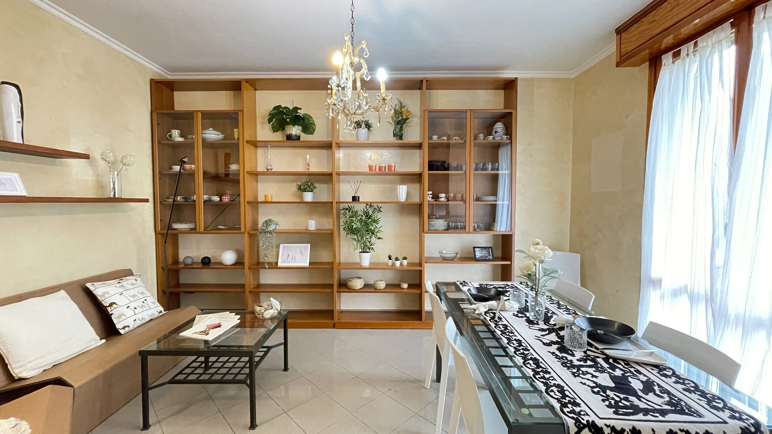Soggiorno moderno con divano marrone, tavolino in vetro, libreria in legno con decorazioni e piante, tavolo da pranzo con tovaglia bianca e nera, sedie bianche e lampade a sospensione.