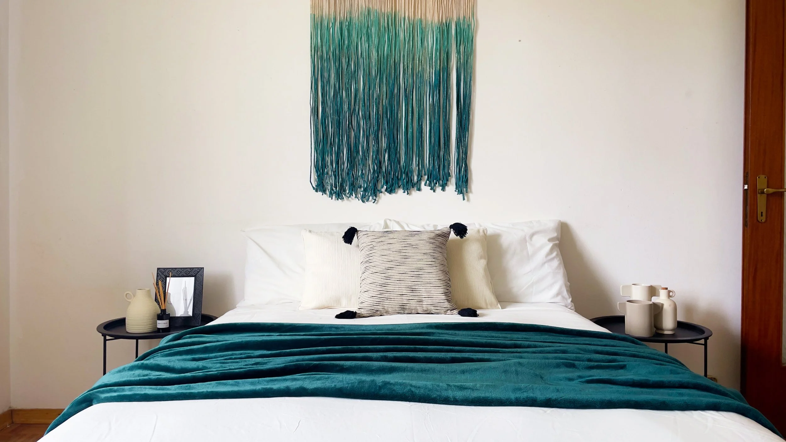 Camera da letto semplice e moderna con letto matrimoniale, coperta verde, cuscini decorativi, tappeto in stile boho appeso alla parete, e comodini con accessori.
