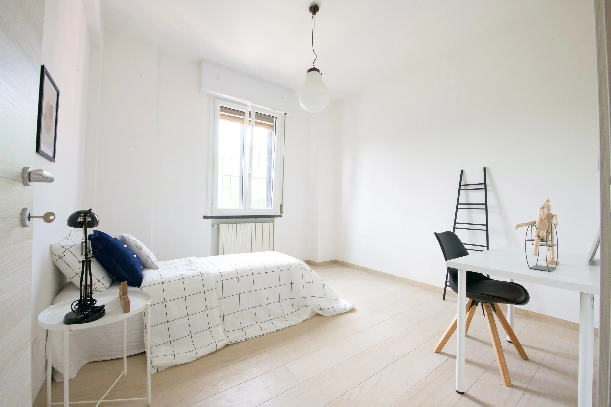 Camera da letto minimalista con letto singolo, coperta a quadri bianchi e neri, tavolino rotondo bianco, lampada, scrivania bianca con sedia nera e decorazione in legno, finestra e pareti bianche, pavimento in legno chiaro.