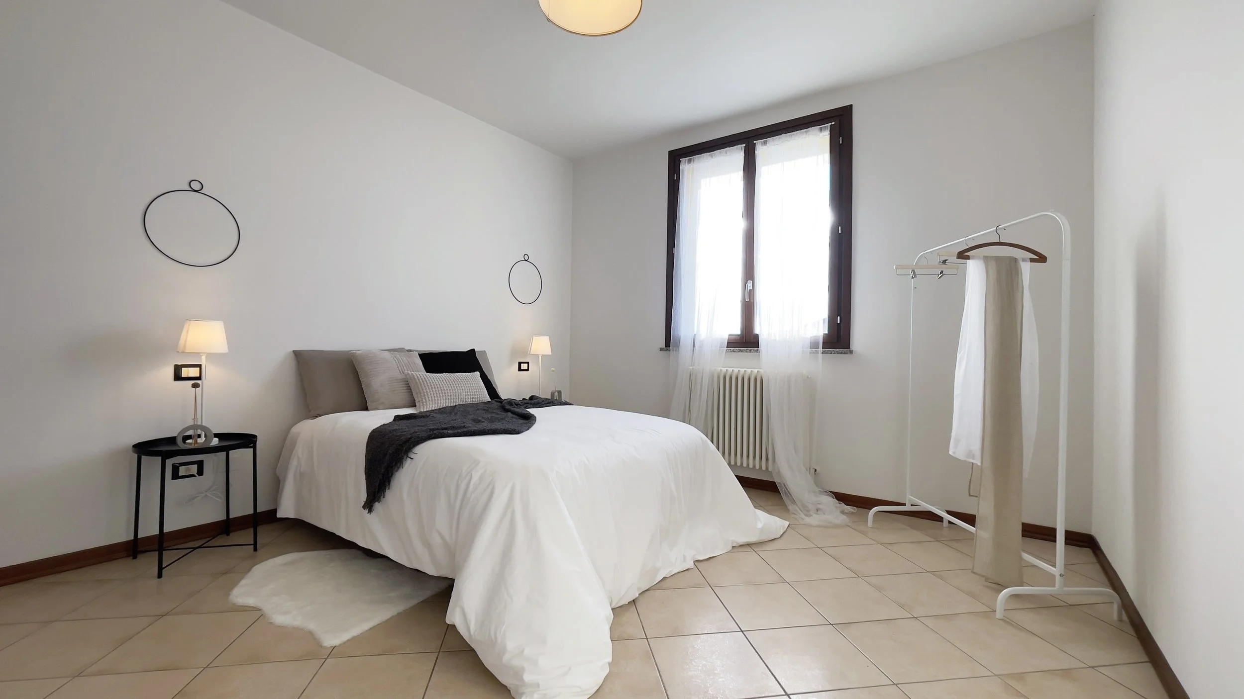 Camera da letto moderna con letto matrimoniale bianco, cuscini grigi, lampade da comodino, appendiabiti e finestra con tende leggere.