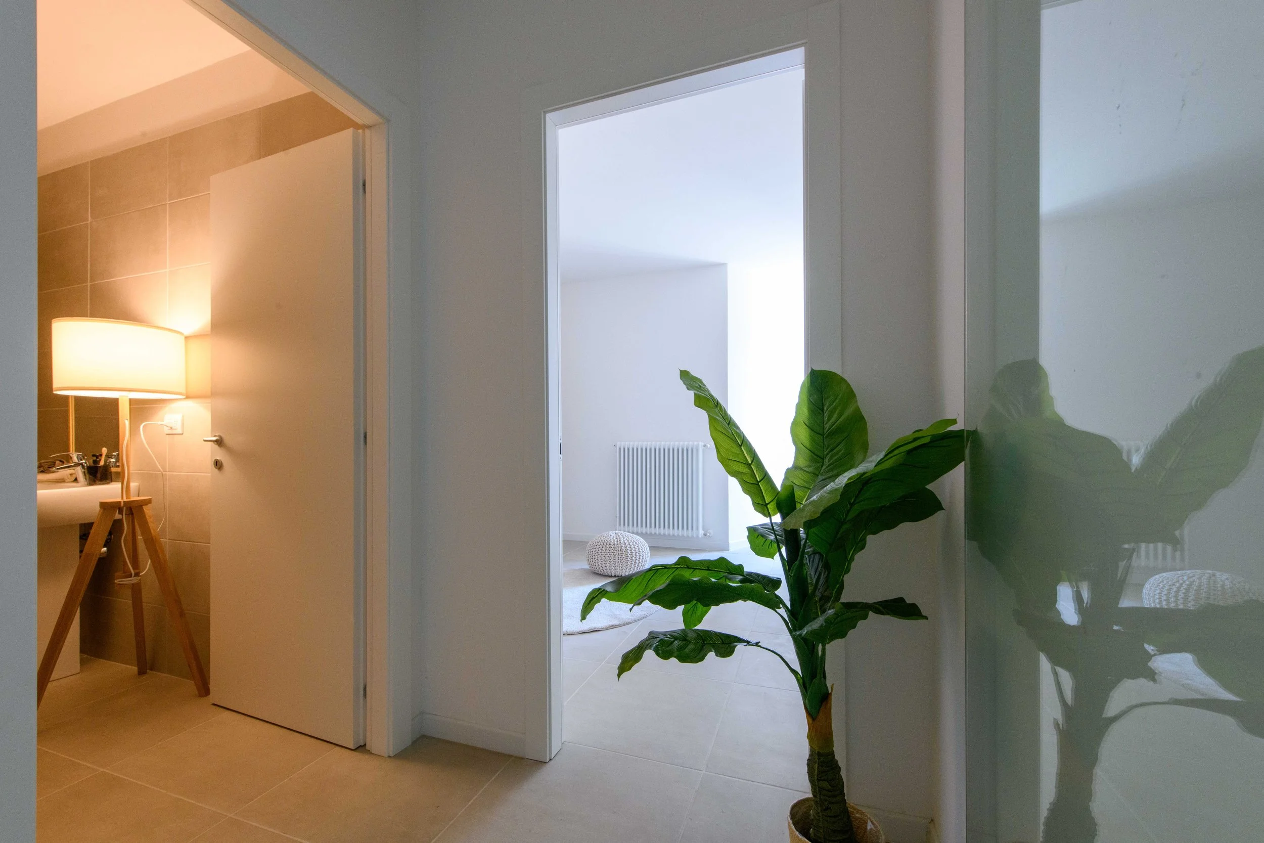 Corridoio di un appartamento moderno con pianta in vaso, lampada accesa in bagno a sinistra, e stanza luminosa a destra con radiatore e pouf.