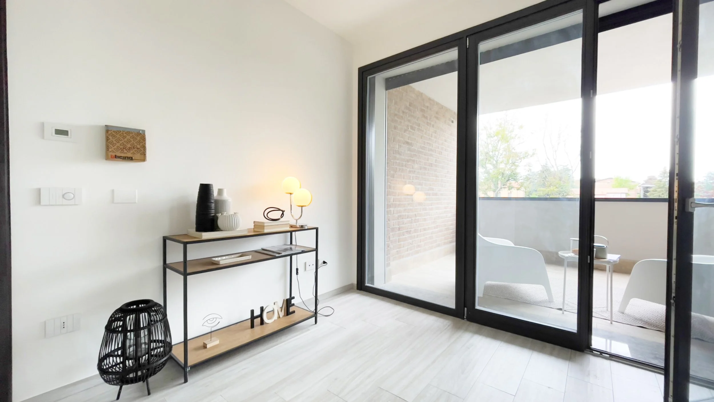 Interno di una stanza moderna con pavimento in legno chiaro, una console con lampada, vasi e decorazioni, accanto a una grande finestra con porta scorrevole che dà su un balcone con sedie bianche.