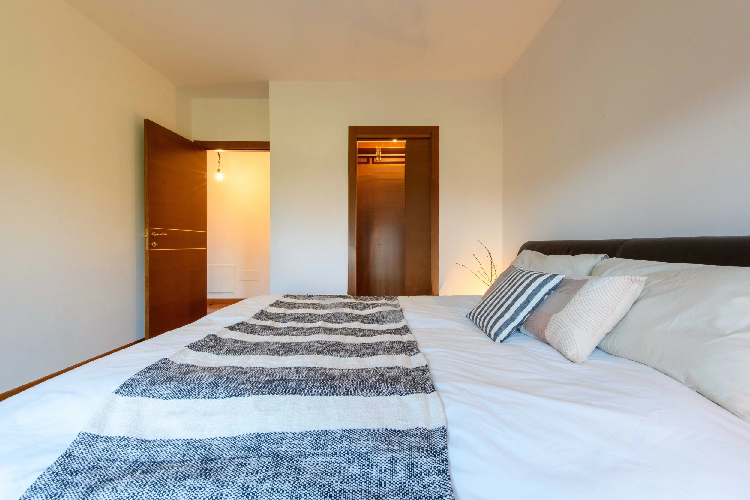 Camera da letto moderna con letto matrimoniale, coperta a righe, cuscini decorativi, porte in legno aperte, illuminazione soffusa e pareti chiare.