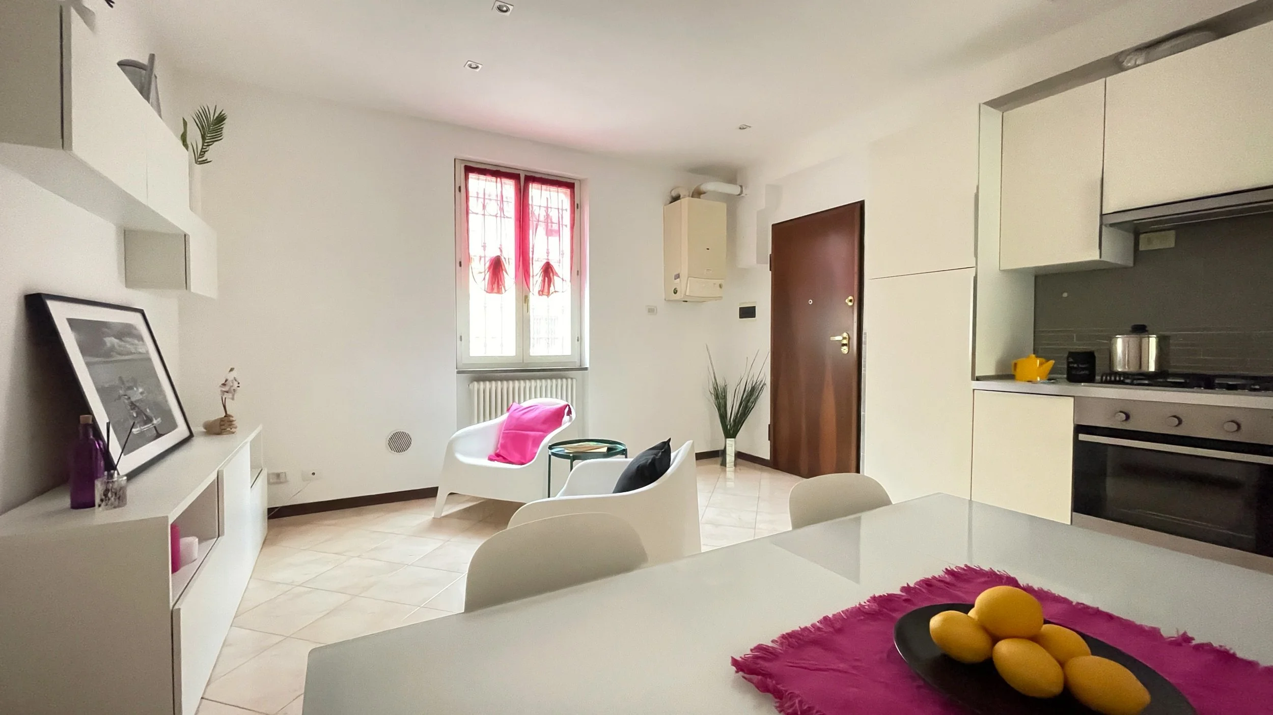 Un soggiorno moderno con cucina integrata. Ci sono sedie bianche moderne, un tavolo con un panno rosa e limoni su un vassoio. Una finestra con tende rosa, mobili bianchi e stoviglie sulla cucina completano l'ambiente.