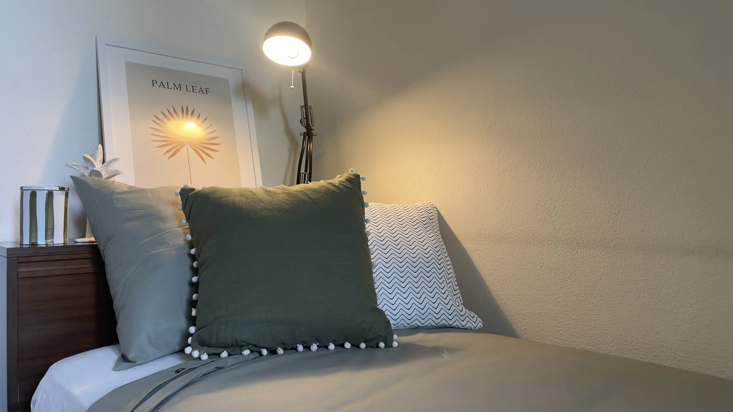 Camera da letto accogliente con letto, cuscini verdi e bianchi, lampada da tavolo, cornice con grafica "Palm Leaf" e decorazione a forma di foglia su un comodino.