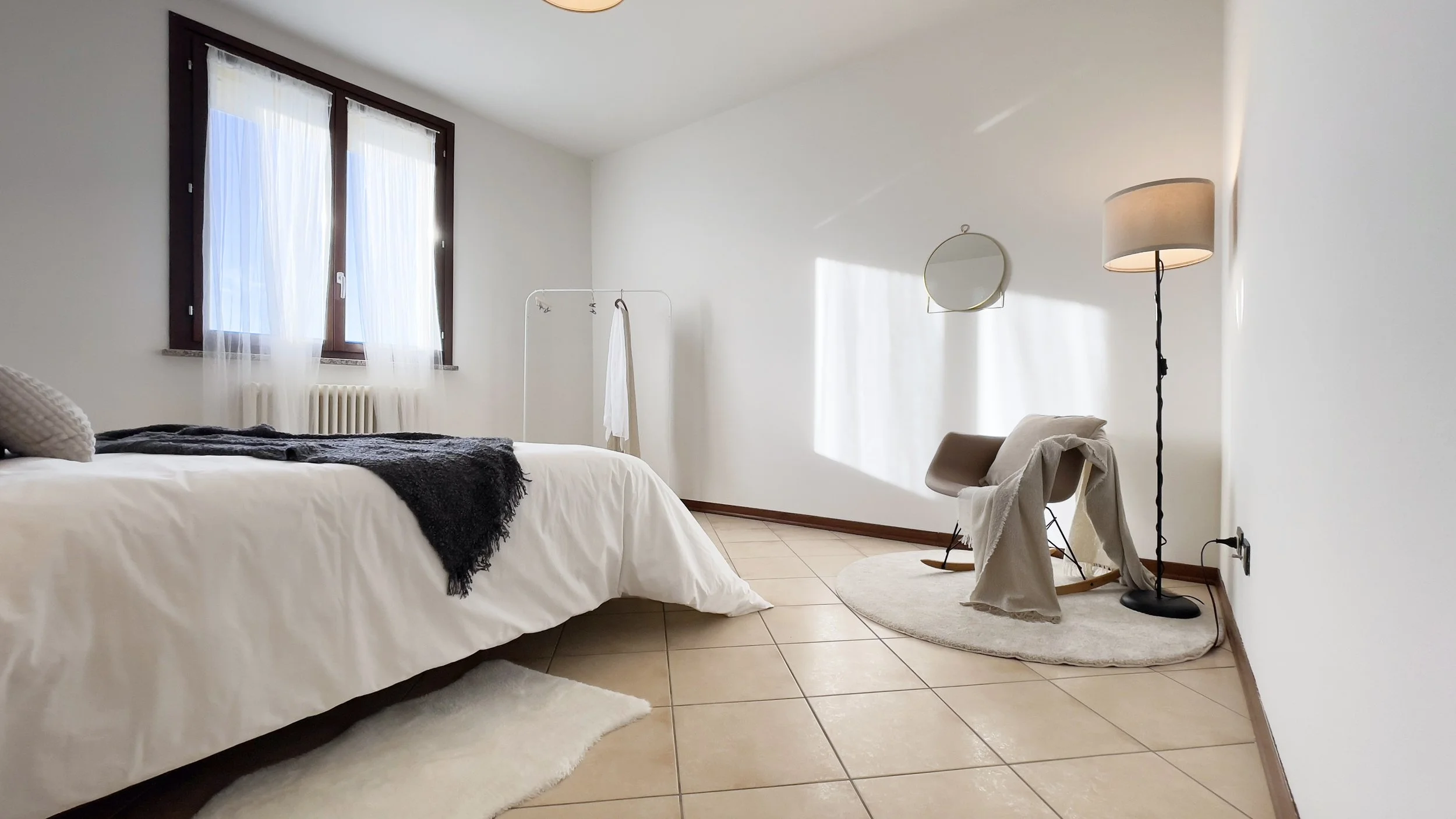 Camera da letto minimalista con letto, sedia, lampada e finestra con tende bianche.