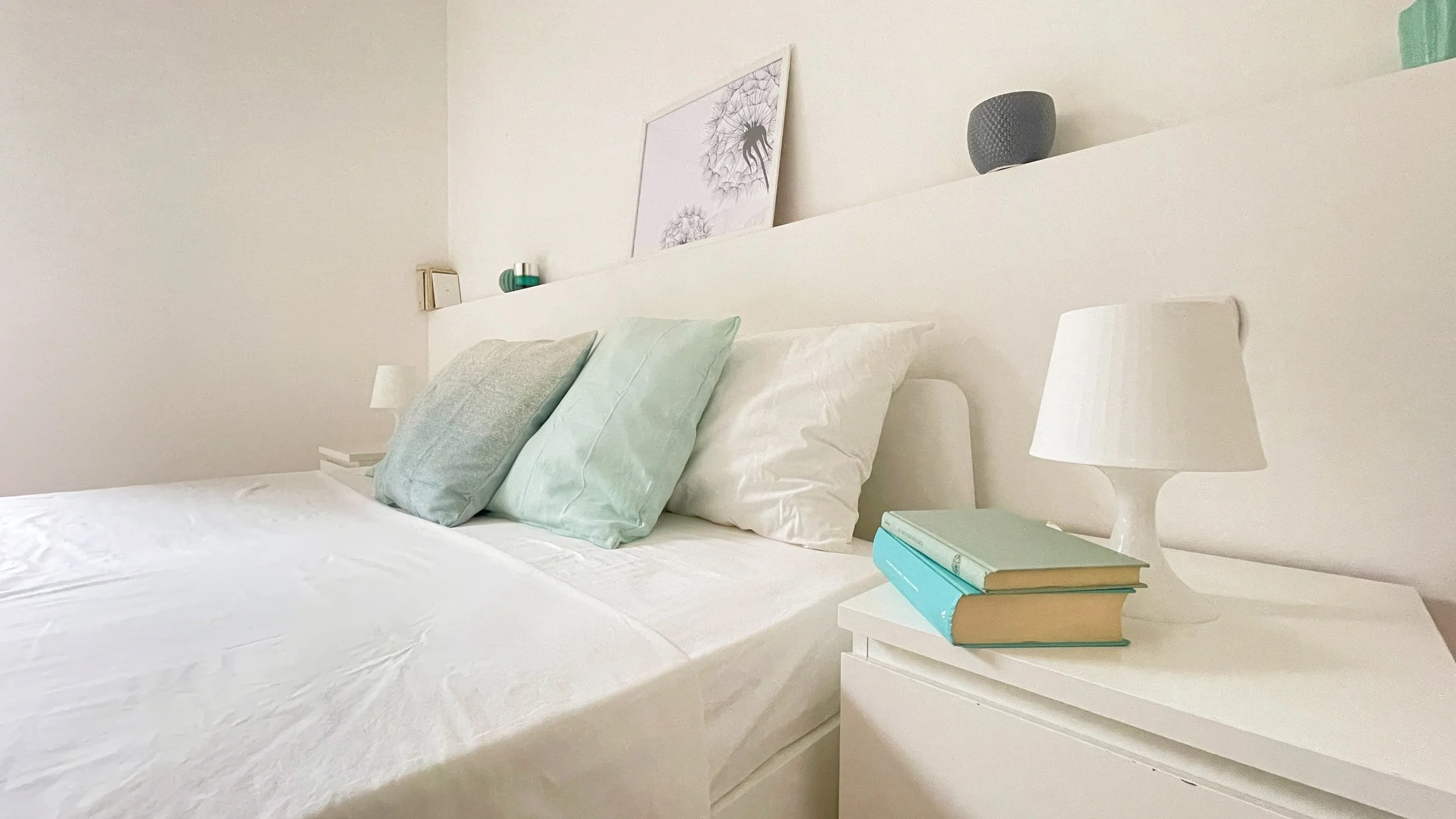 Camera da letto con letto matrimoniale, cuscini bianchi e verde acqua, lampada da comodino bianca e libri su un comodino bianco, decorazione da parete con fiori stilizzati.