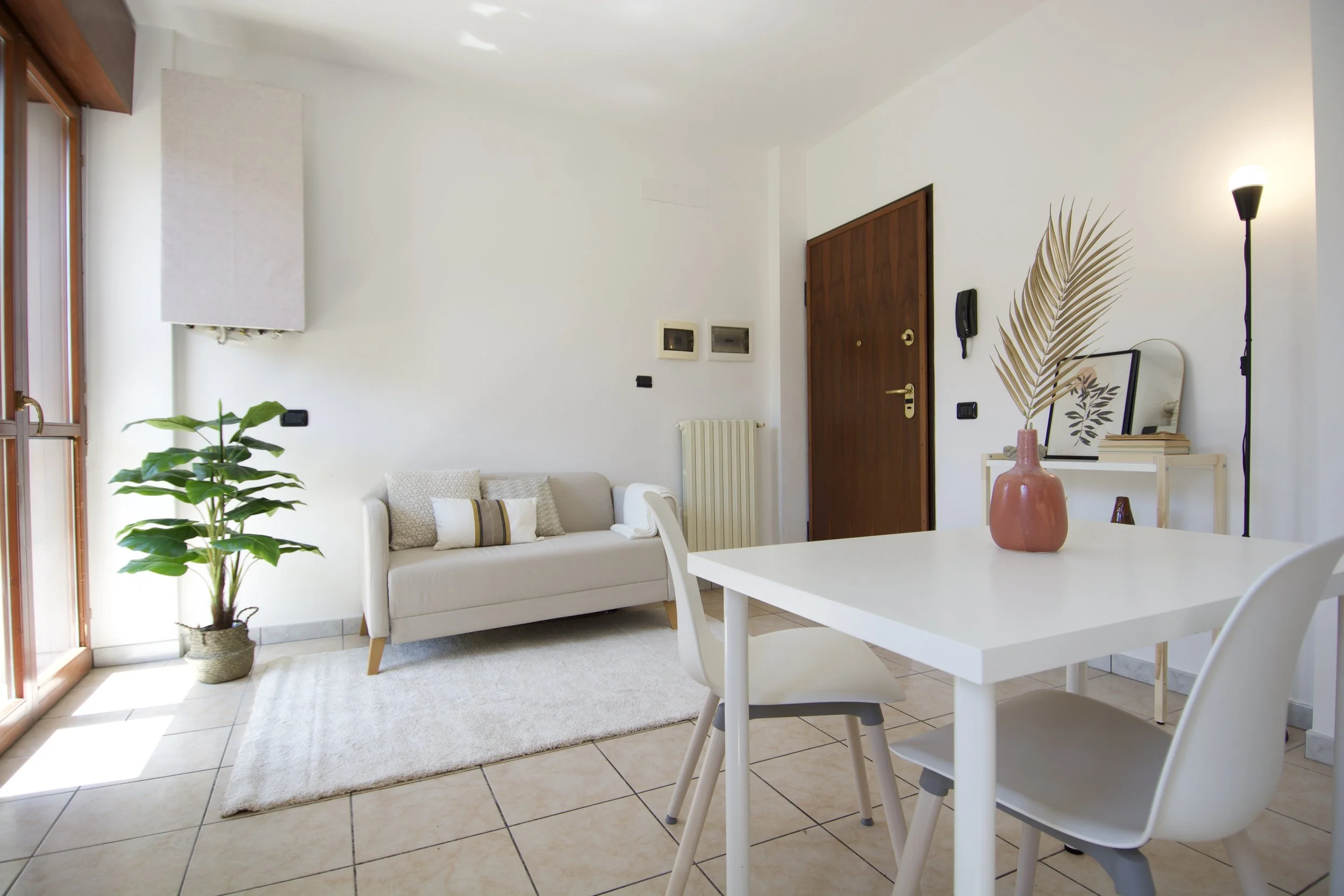 Soggiorno moderno e luminoso con divano beige, pianta decorativa, tavolo con sedie bianche, vaso con decorazione, porta in legno, e pavimento piastrellato.