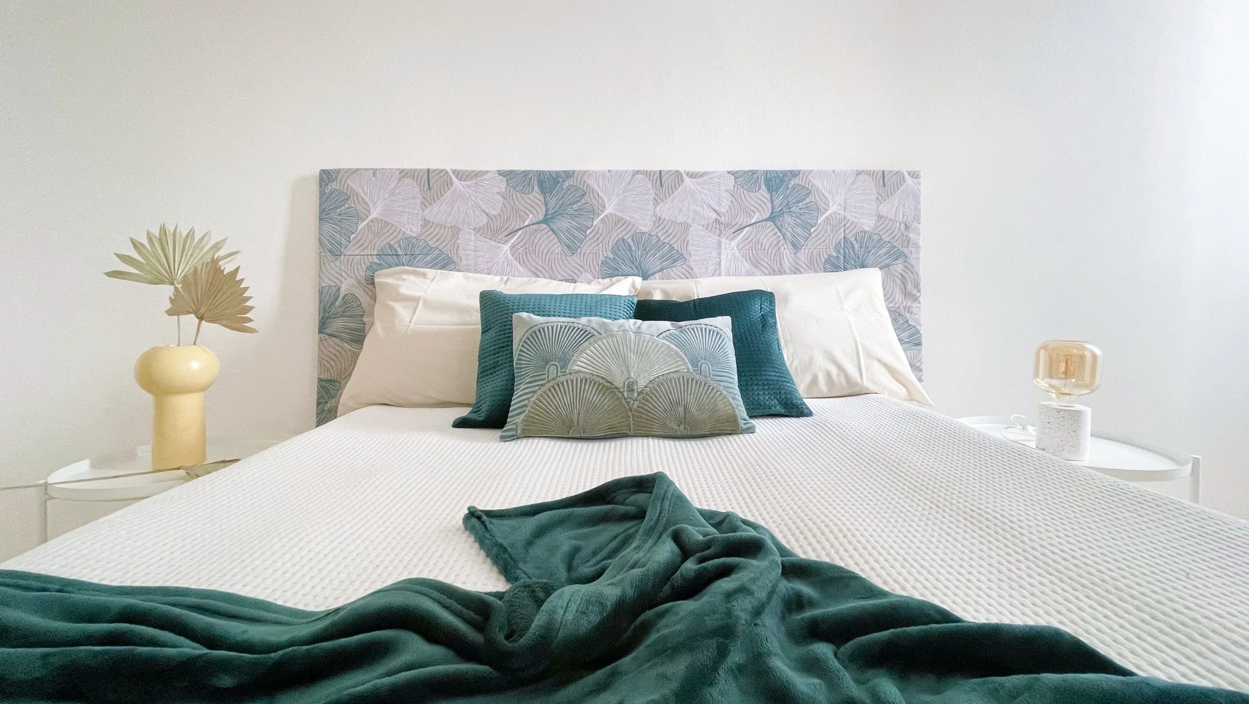 Camera da letto moderna con letto matrimoniale, cuscini decorativi blu e verde, testiera con motivo a foglie, coperta verde scuro, comodini con lampada e vaso decorativo.