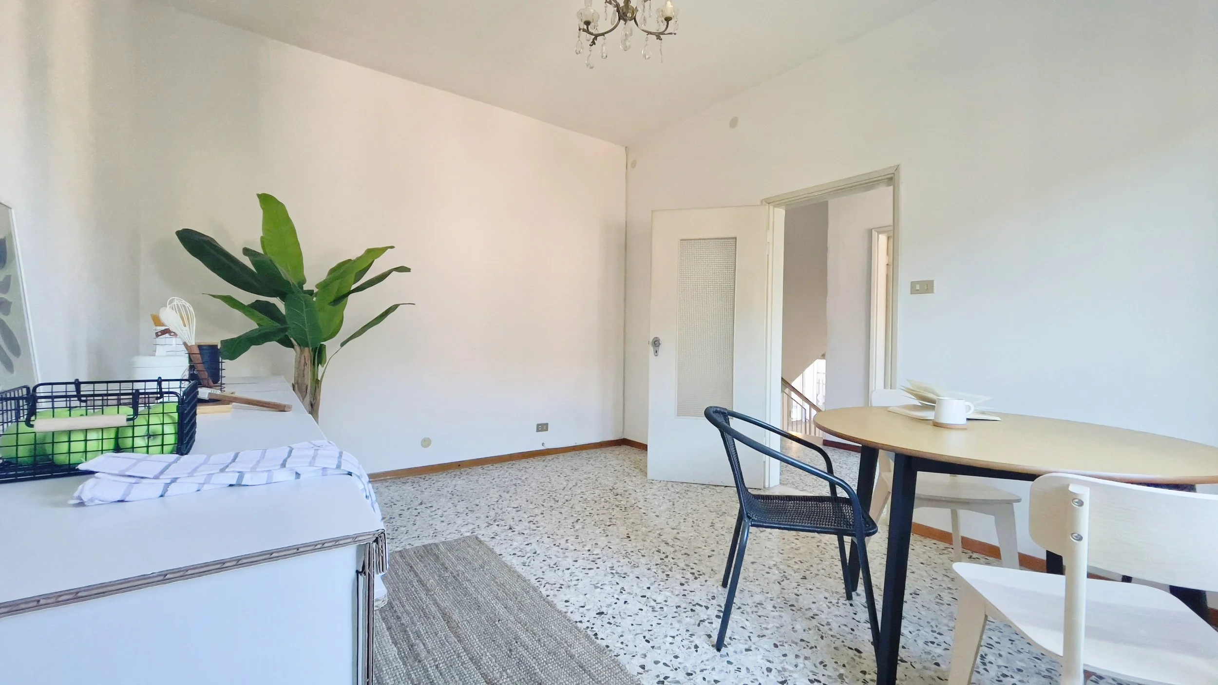 Stanza minimalista con tavolo rotondo e sedie, una pianta verde in un angolo, e un piccolo lampadario al soffitto. Il pavimento è in marmo e c'è un cesto con mele verdi sul mobile.