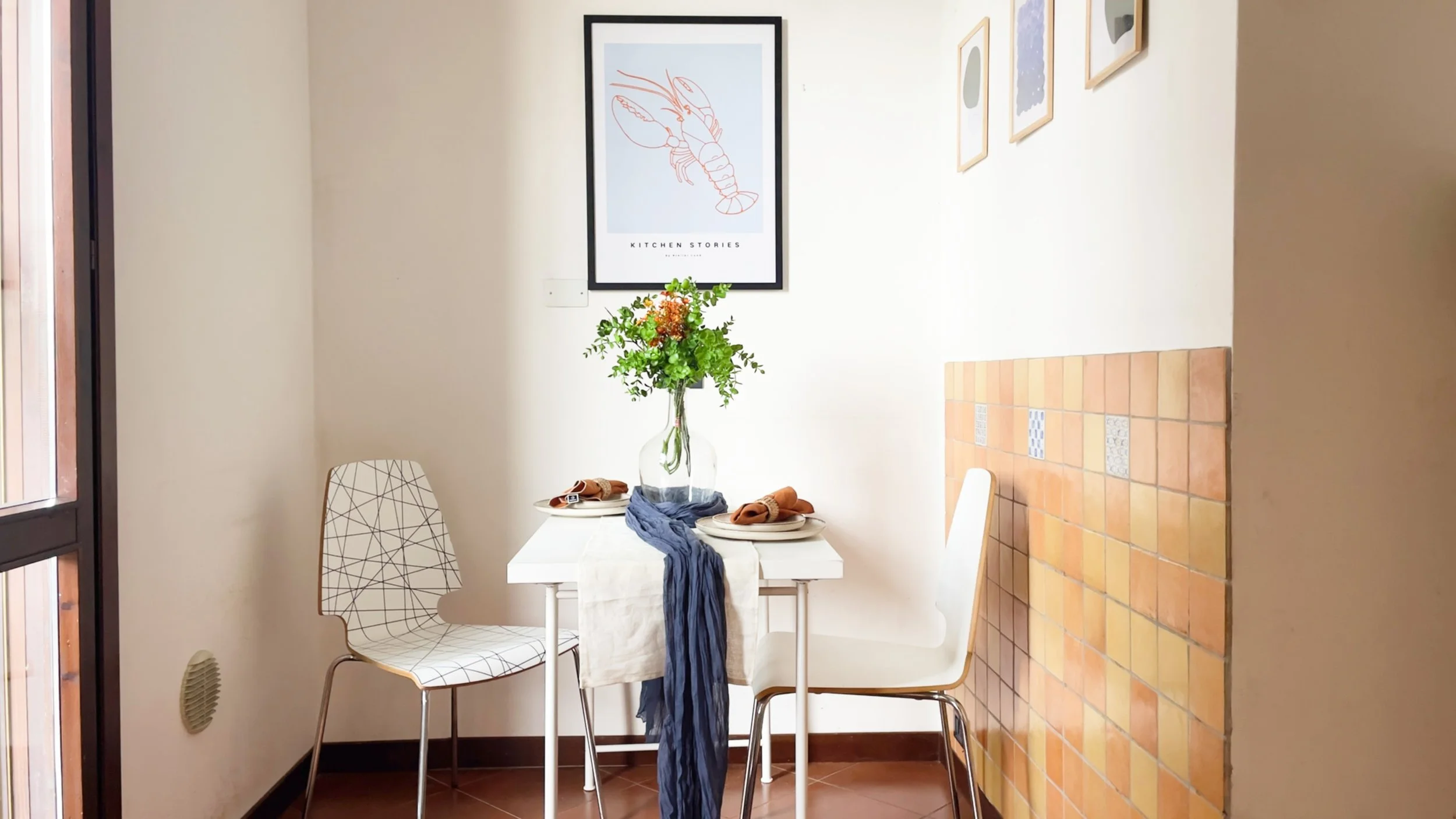 Angolo pranzo con tavolino bianco, due sedie moderne, e decorazioni come un vaso di fiori e tovaglioli arancioni. Sulla parete ci sono quadri e piastrelle colorate.