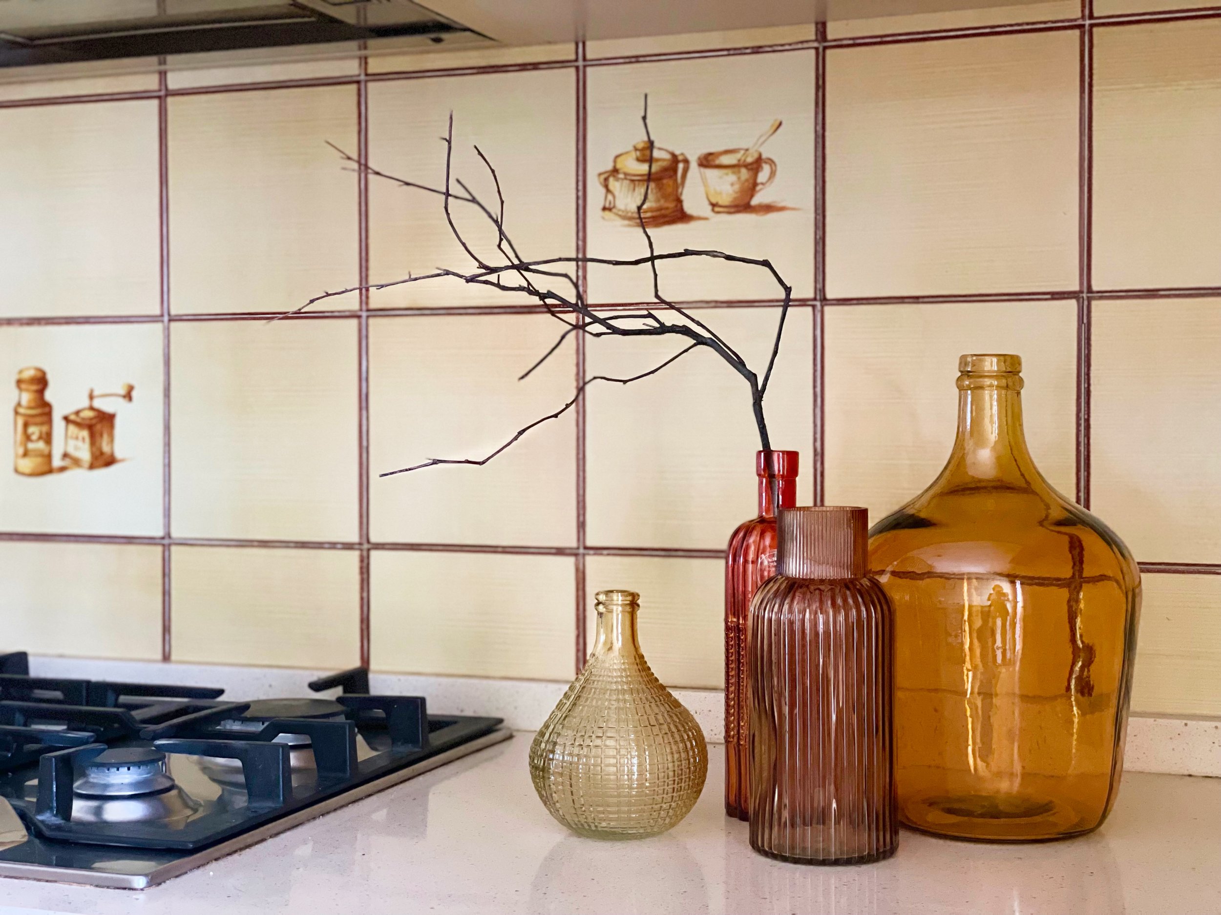 Piano cottura con fornelli a gas accanto a tre bottiglie decorative in vetro colorato, una rossa, una marrone e una gialla, su un piano di lavoro con piastrelle beige e motivi di utensili da cucina.