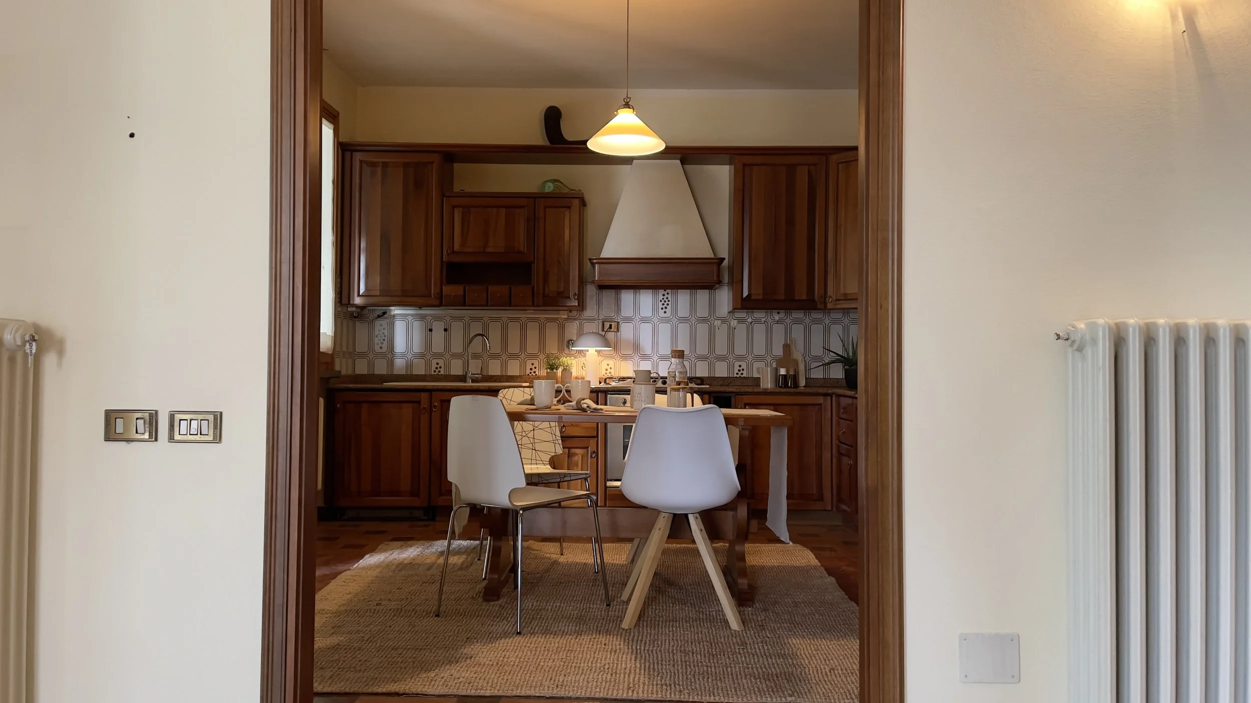 Cucina con mobili in legno, tavolo con sedie bianche, lampada a sospensione, piano di lavoro con utensili e oggetti vari.