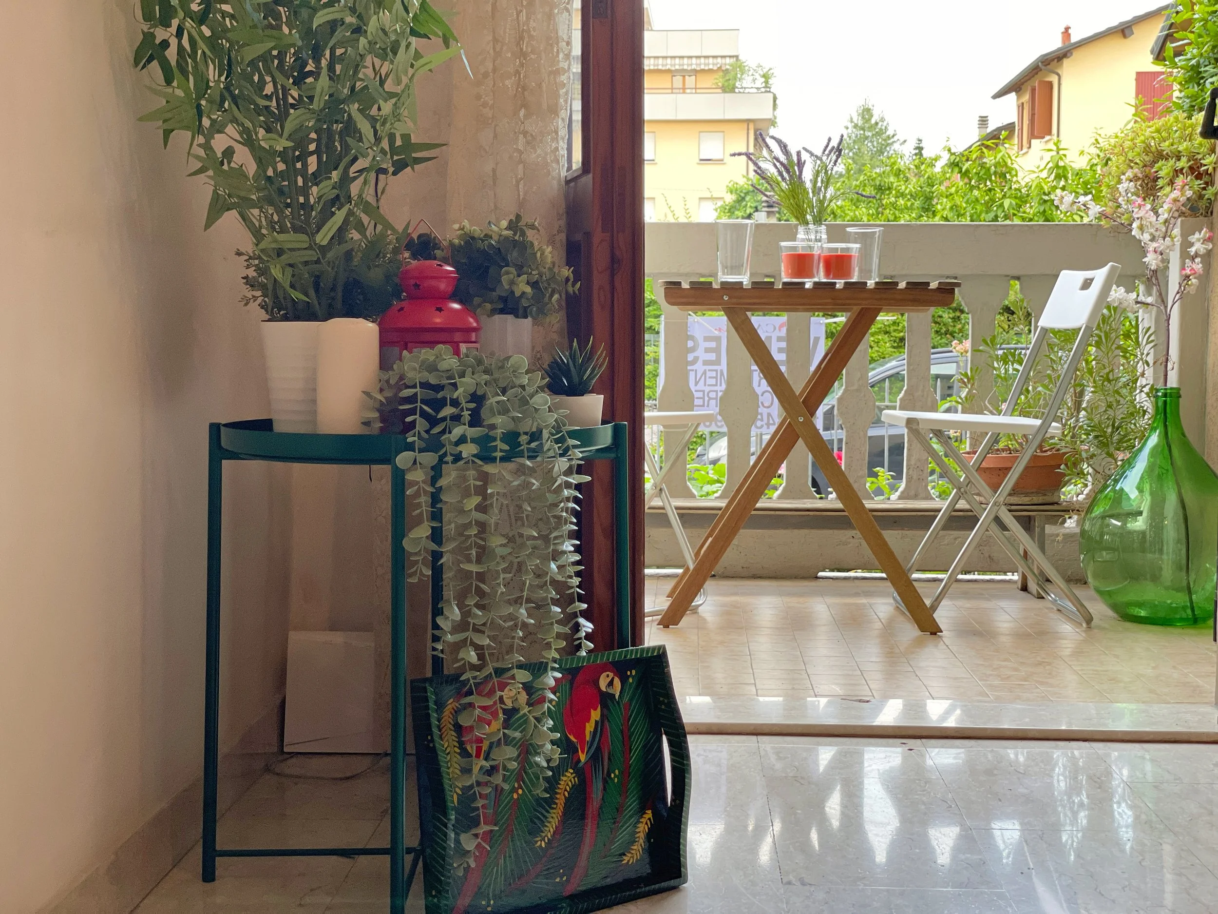 Interno di un appartamento con porta aperta su un balcone. Vicino alla porta c'è un tavolino verde con piante e una lanterna rossa. Sul balcone ci sono un tavolo di legno e due sedie bianche pieghevoli, e una grande bottiglia verde. Ci sono piante e 