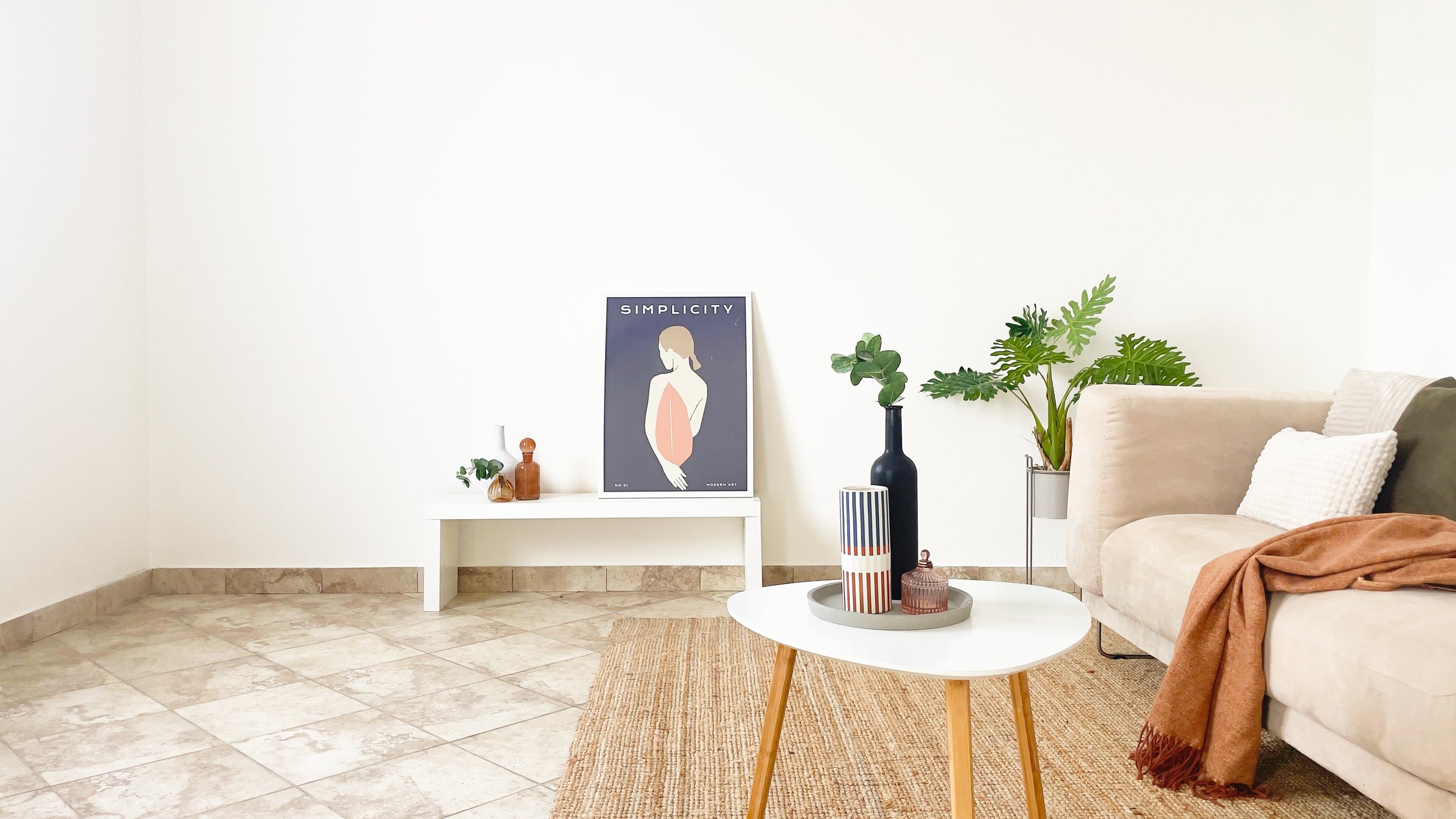 Soggiorno minimalista con divano beige, tavolino bianco con decorazioni, piante e un poster d'arte moderna con scritta "Simplicity". La stanza ha pavimento in piastrelle chiare.