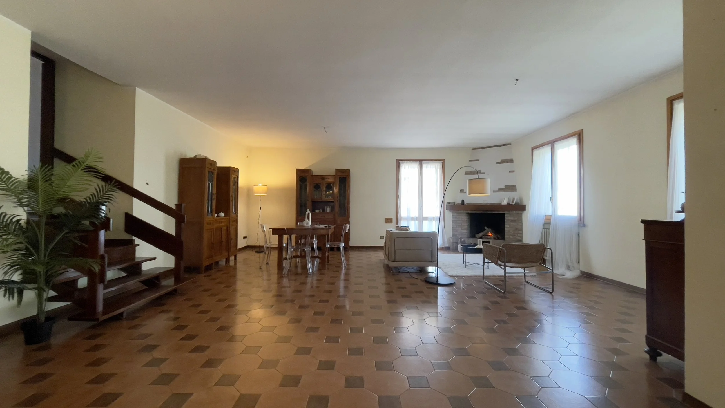 Soggiorno spazioso con pavimento in piastrelle esagonali marroni, arredato con un divano beige, un tavolo in legno con sedie trasparenti, credenze in legno, un camino e una lampada ad arco. A sinistra, si nota una scala in legno e una pianta decorati