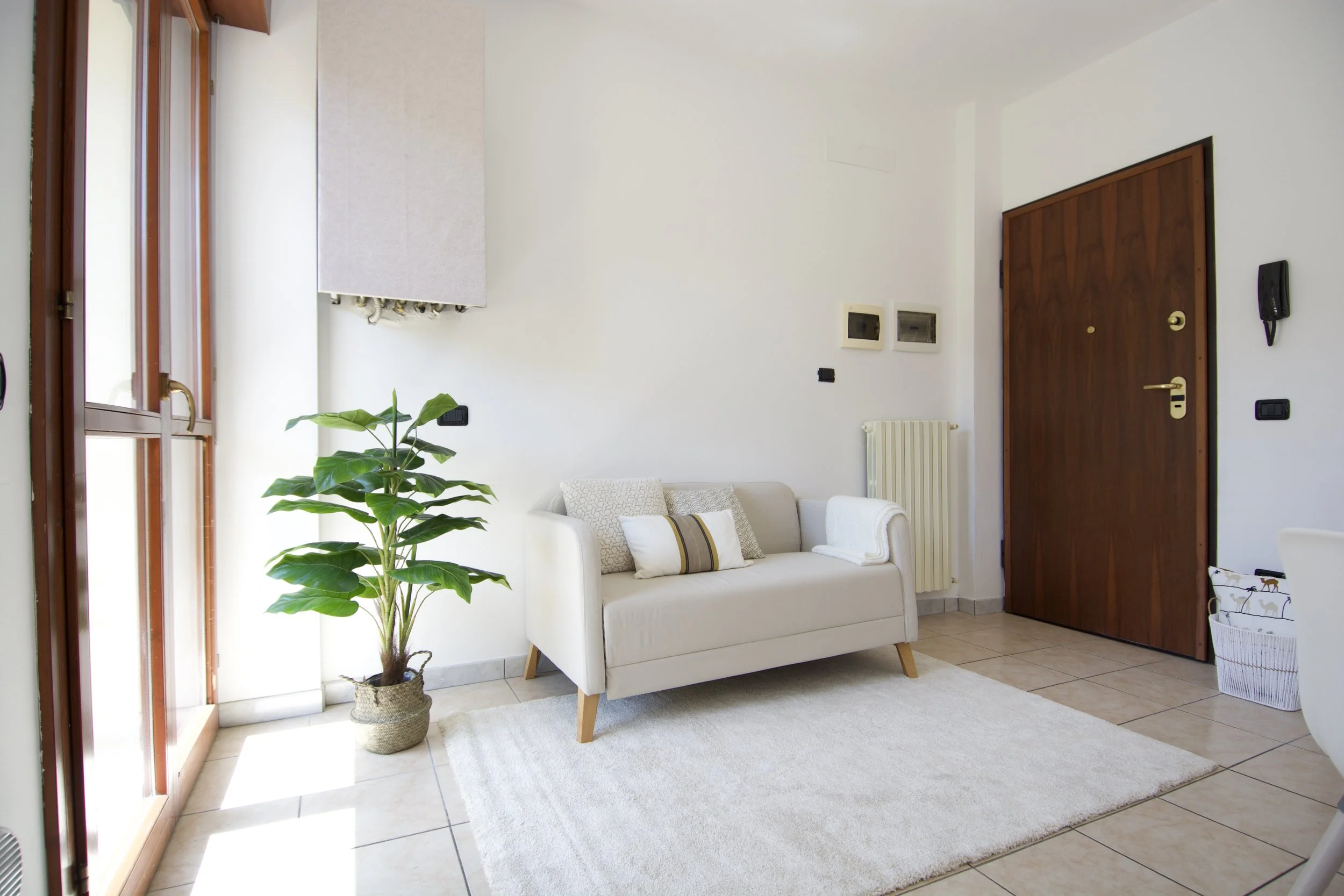 Interno di un soggiorno minimalista con divano bianco e pianta decorativa accanto a una finestra luminosa. Porta in legno e radiatore a parete visibili.
