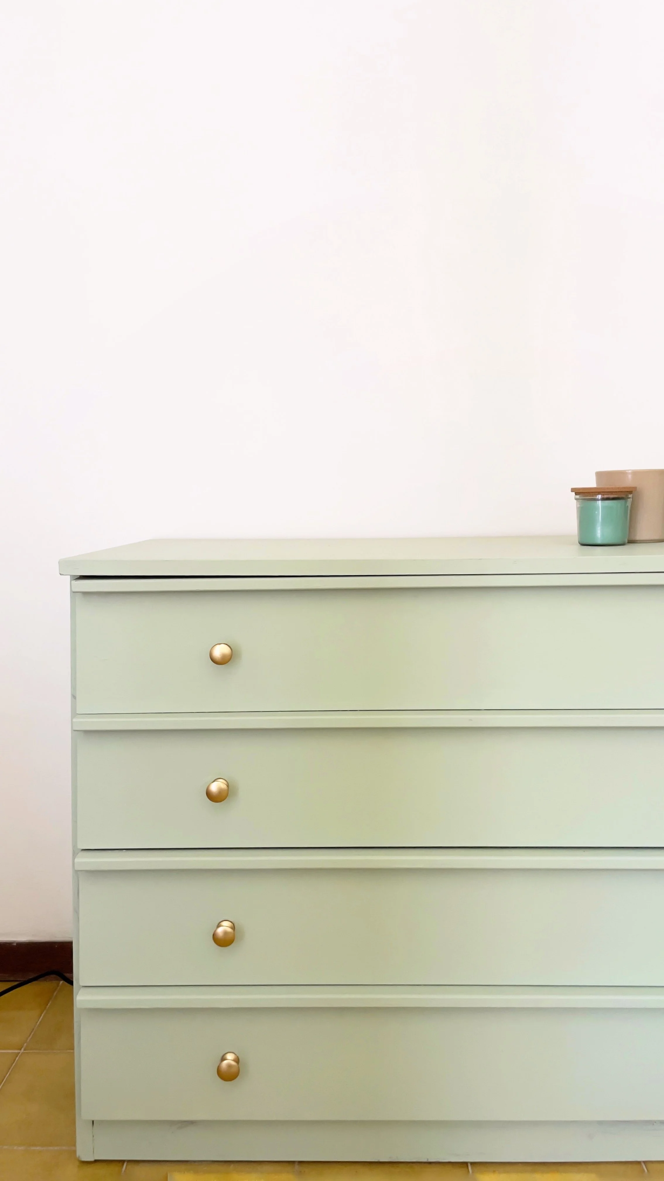 Cassettiera verde chiaro con pomelli dorati, decorata con vasetti sulla superficie superiore.