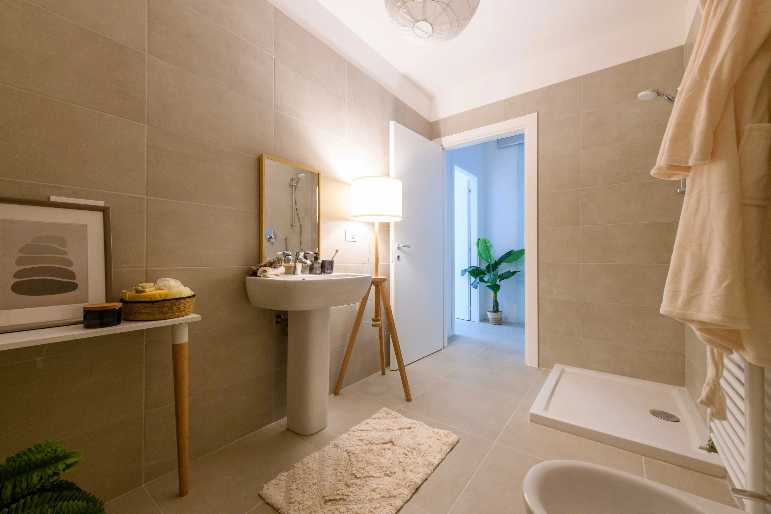 Bagno moderno con lavabo, specchio, doccia, lampada da terra, pianta decorativa e opere d'arte su un tavolino. Pareti e pavimento con piastrelle beige, accappatoio appeso.