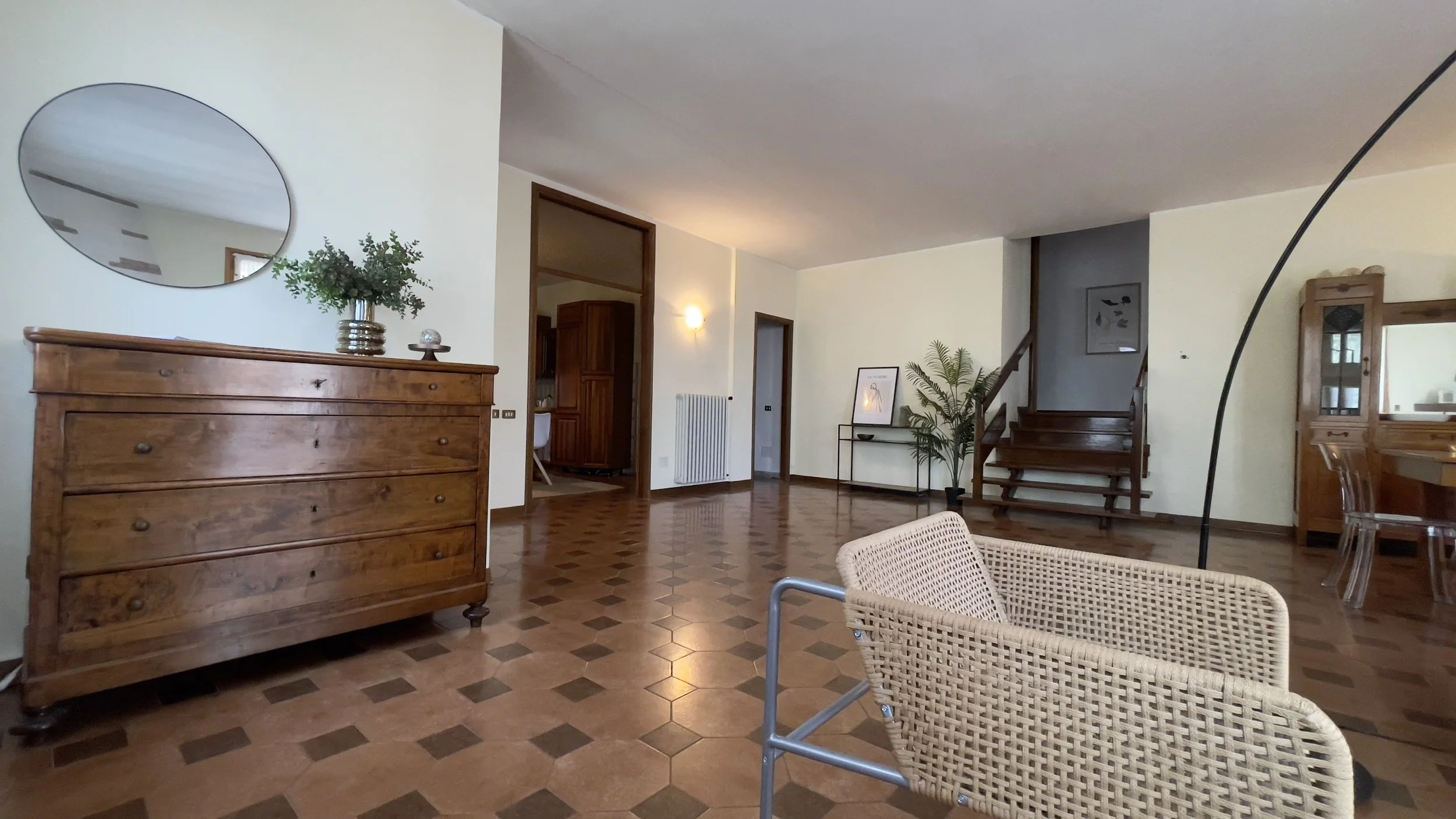 Interno di una stanza spaziosa con un comò in legno scuro, specchio circolare sopra, sedia in vimini e scala in legno sullo sfondo. Decorazioni includono piante e quadri sulle pareti beige.