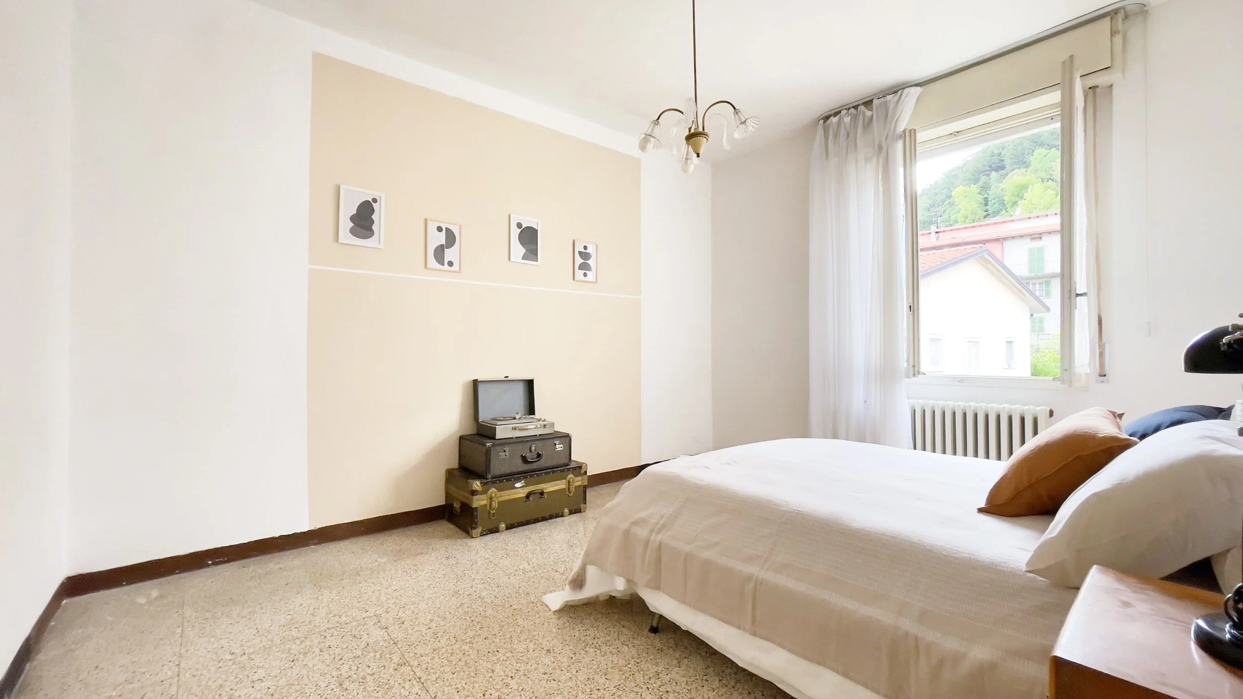 Camera da letto luminosa con letto matrimoniale, tende bianche e finestra aperta. Valigie vintage e giradischi in un angolo, con quadri astratti sulla parete beige. Vista di un edificio e verde fuori.