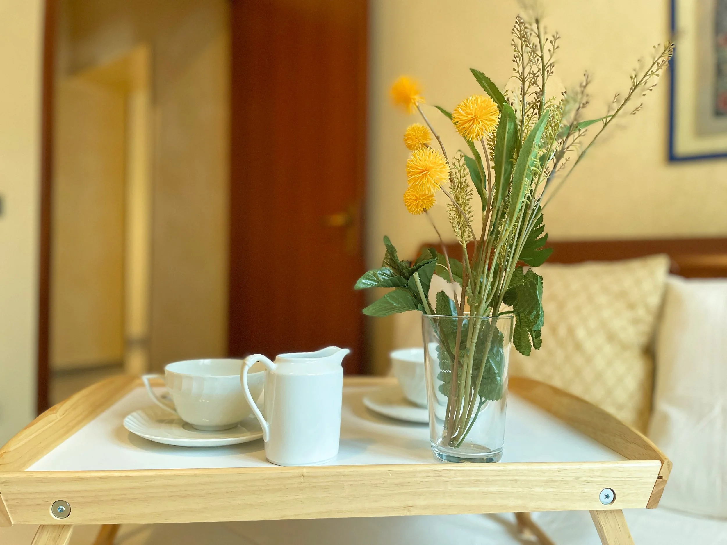 Un vassoio da colazione con una tazza bianca, una brocca e un bicchiere con fiori gialli, su un letto con cuscini chiari.