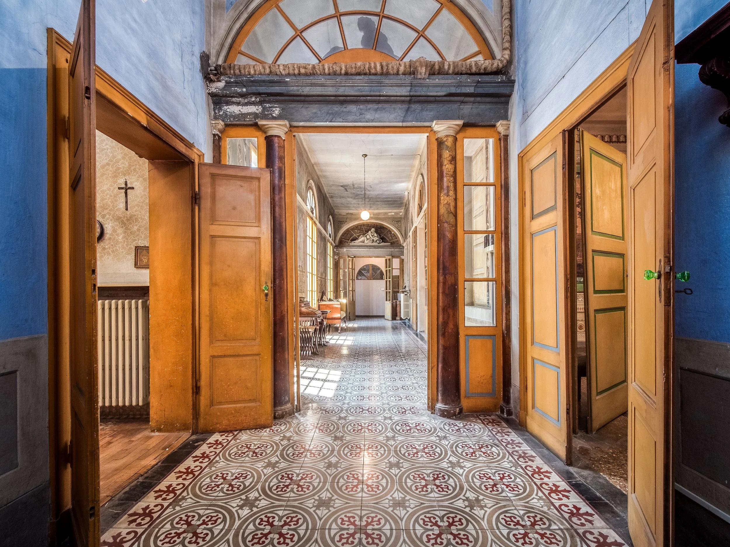 Corridoio di un edificio storico con pavimento piastrellato decorato, porte in legno aperte e pareti blu e beige.