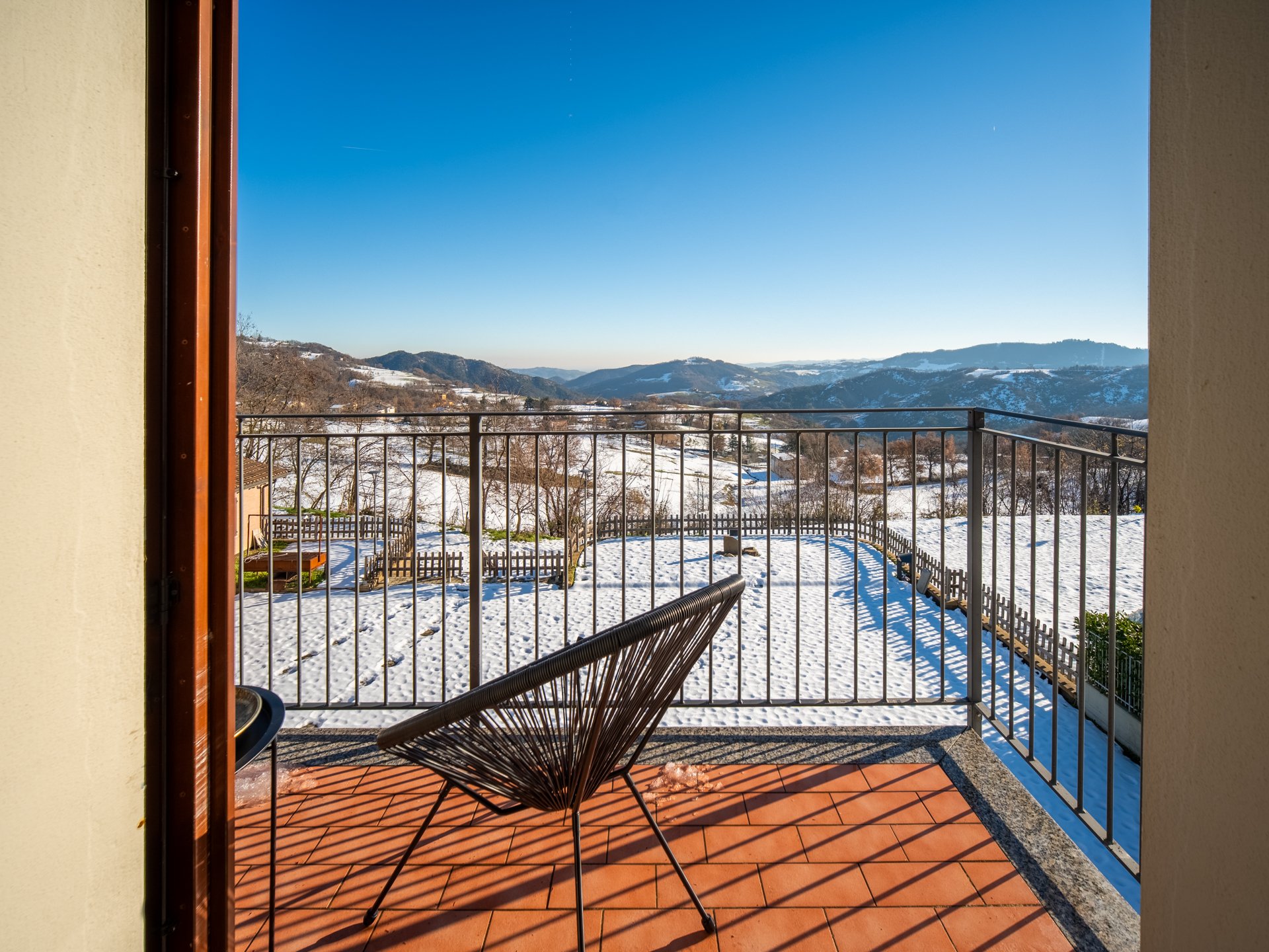 Veduta da un balcone con pavimento in cotto, su un paesaggio invernale innevato con colline in lontananza e cielo sereno, e una sedia in primo piano.
