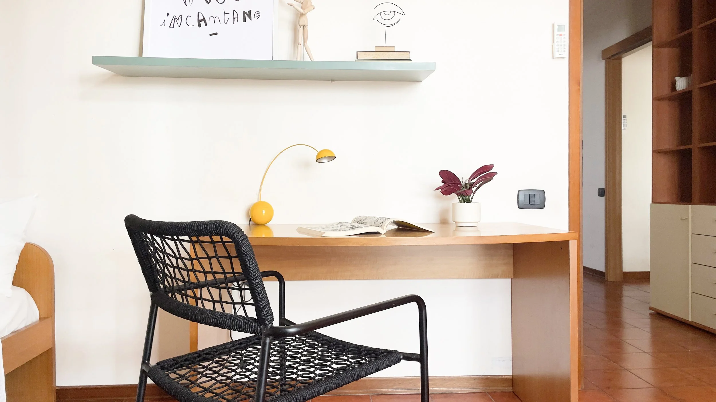 Interno di una stanza moderna con scrivania in legno, sedia nera intrecciata, lampada gialla, pianta in vaso bianco, e mensola con decorazioni."}