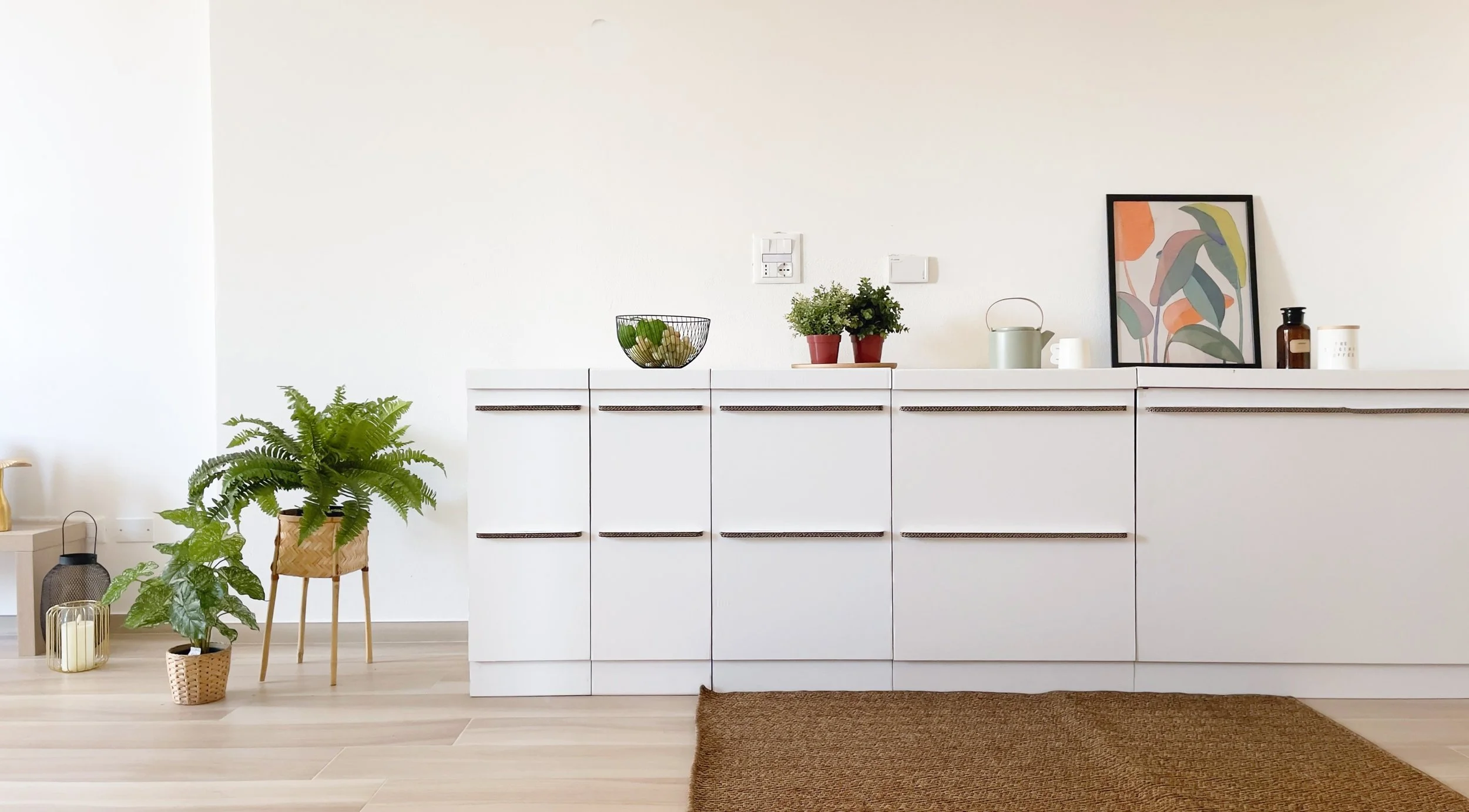Cucina moderna con mobili bianchi, piante verdi in vaso, disegno incorniciato colorato, brocca verde, bottiglia marrone e tappeto marrone su pavimento in legno chiaro.