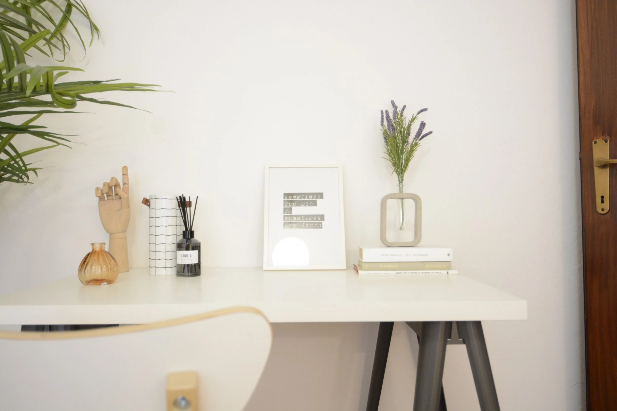 Scrivania bianca con oggetti decorativi, tra cui una mano in legno, un flacone profumato, un vaso con fiori secchi, libri e una cornice con citazione. C'è una pianta a sinistra e una porta a destra.