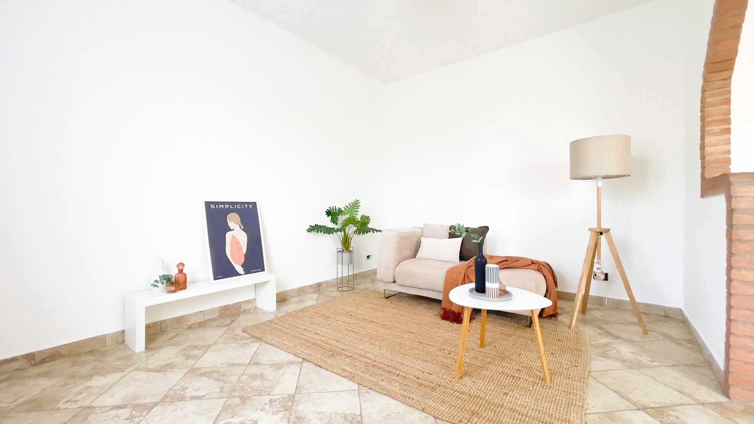 Soggiorno minimalista con divano beige, tavolino rotondo bianco, lampada da terra, piante decorative e poster artistico su una mensola bianca.