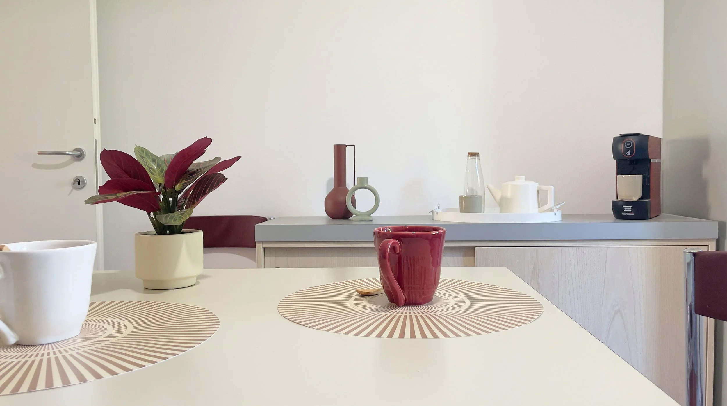 Tavolo con tazze, piantina, placemat e macchina del caffè in una cucina moderna.