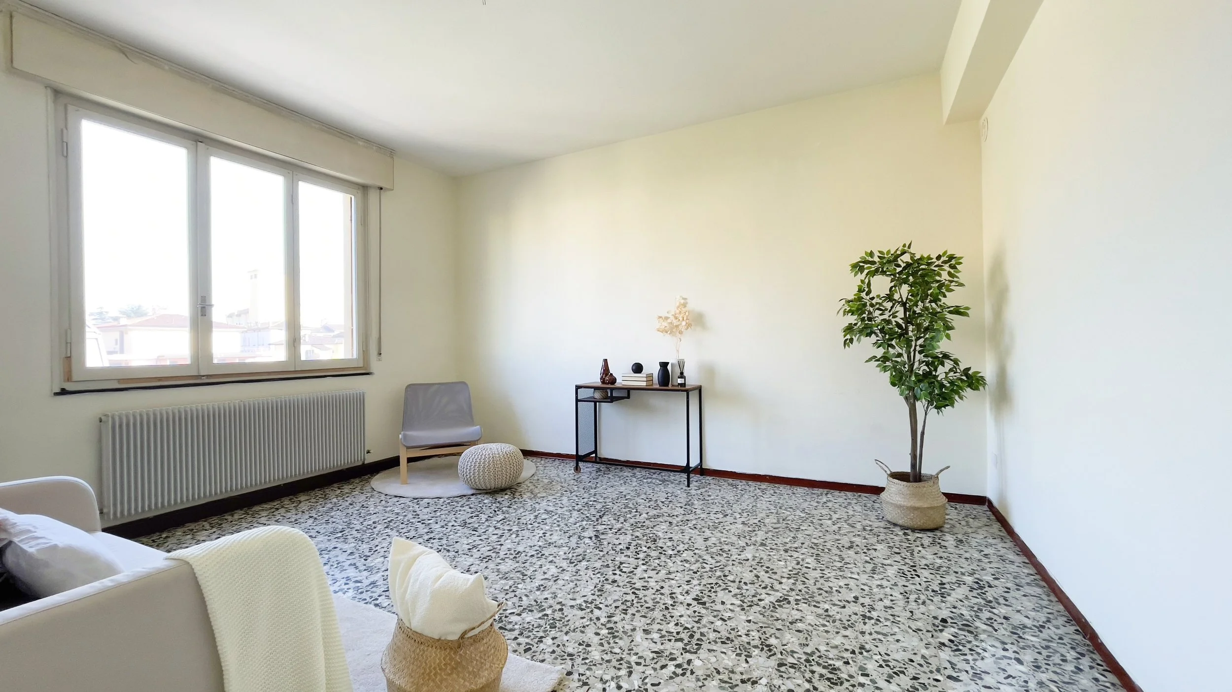 Soggiorno minimalista con pavimento in marmo, finestra grande, poltrona grigia, pouf, tavolino con decorazioni, e pianta in vaso.