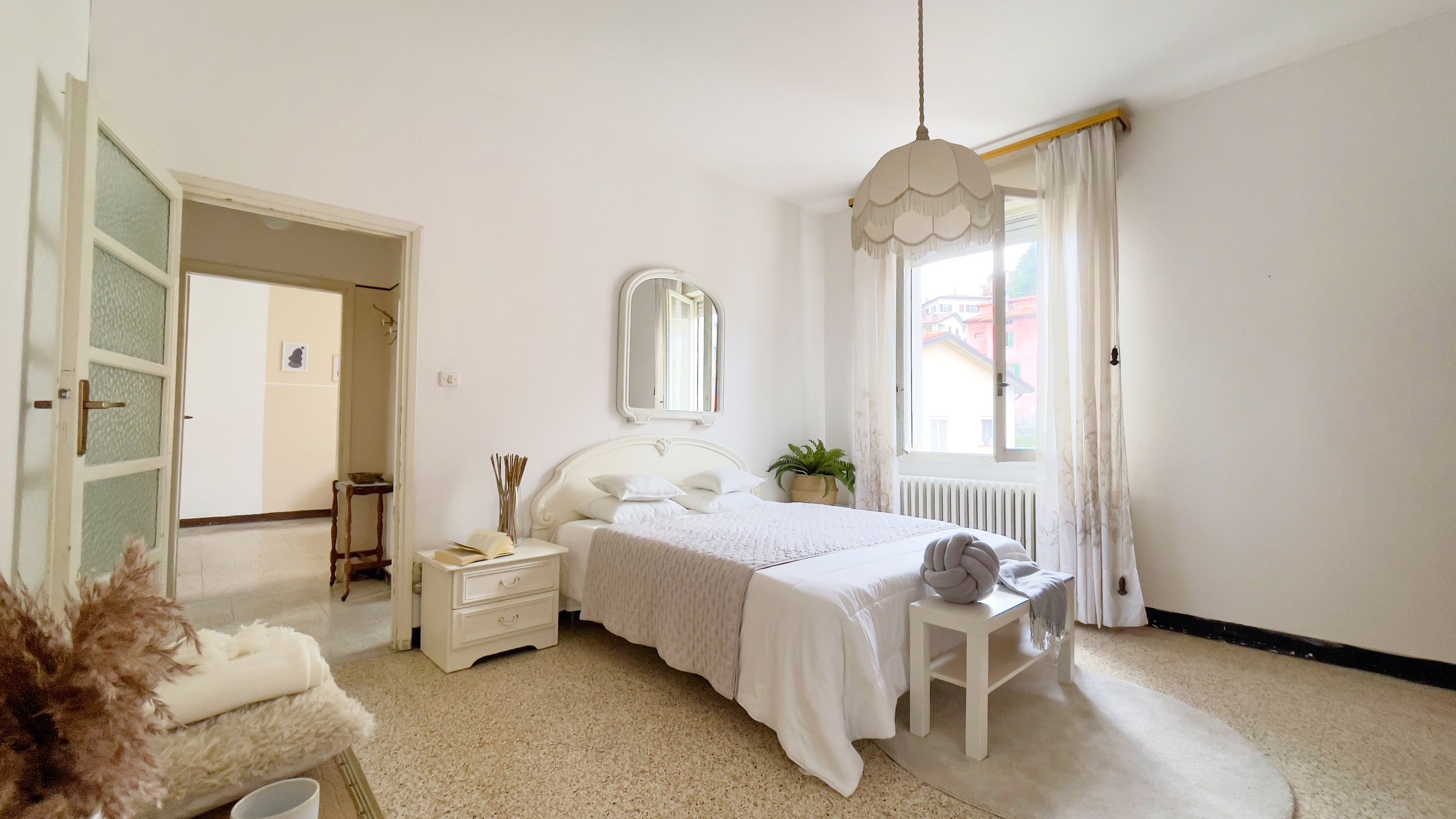 Camera da letto luminosa con decorazioni bianche, letto matrimoniale, comodino, specchio, finestra ampia e lampadario pendente.