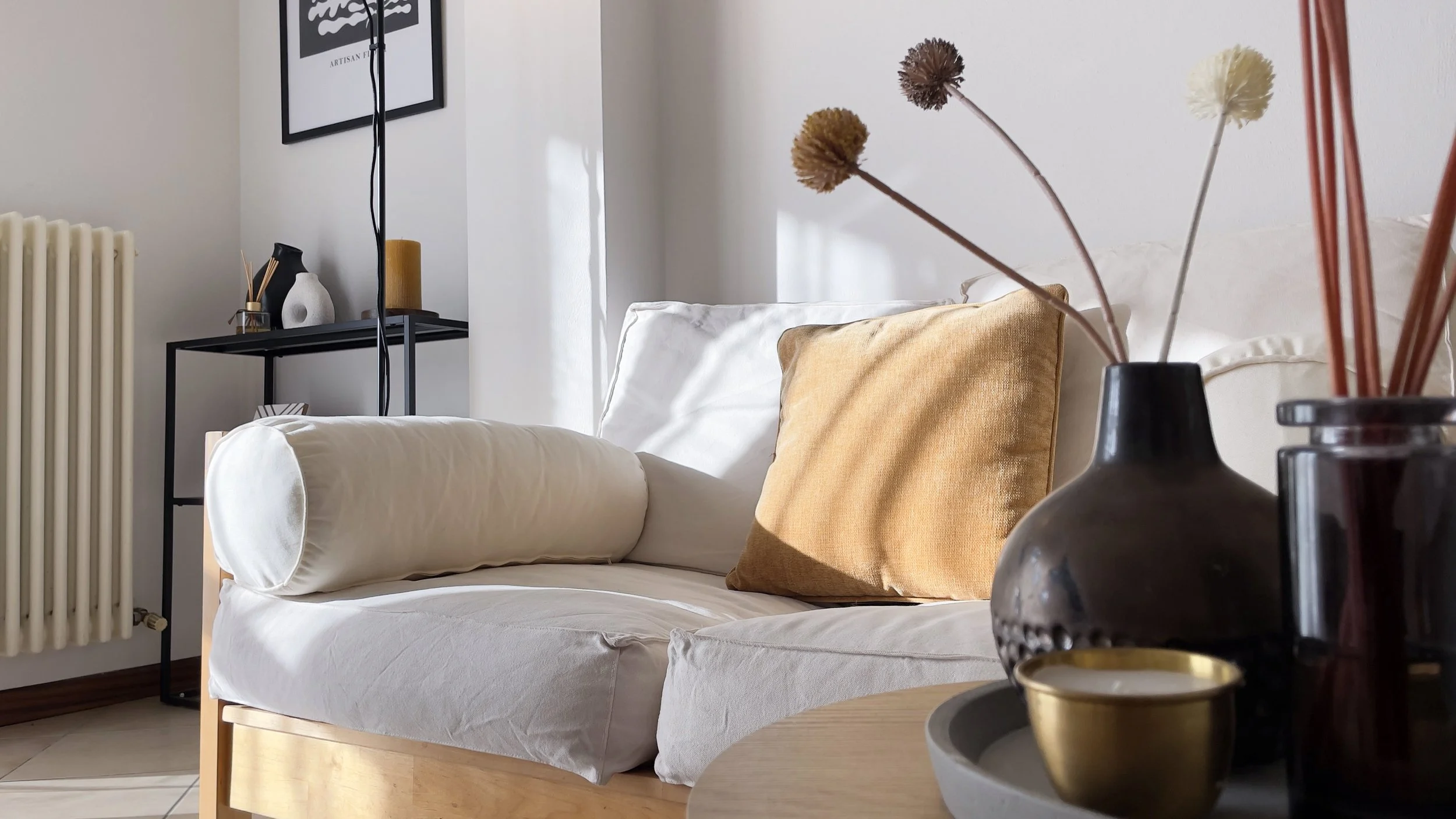 Interno di un salotto moderno, con un divano bianco e cuscini color senape, una mensola nera con decorazioni e una finestra che lascia entrare luce naturale.