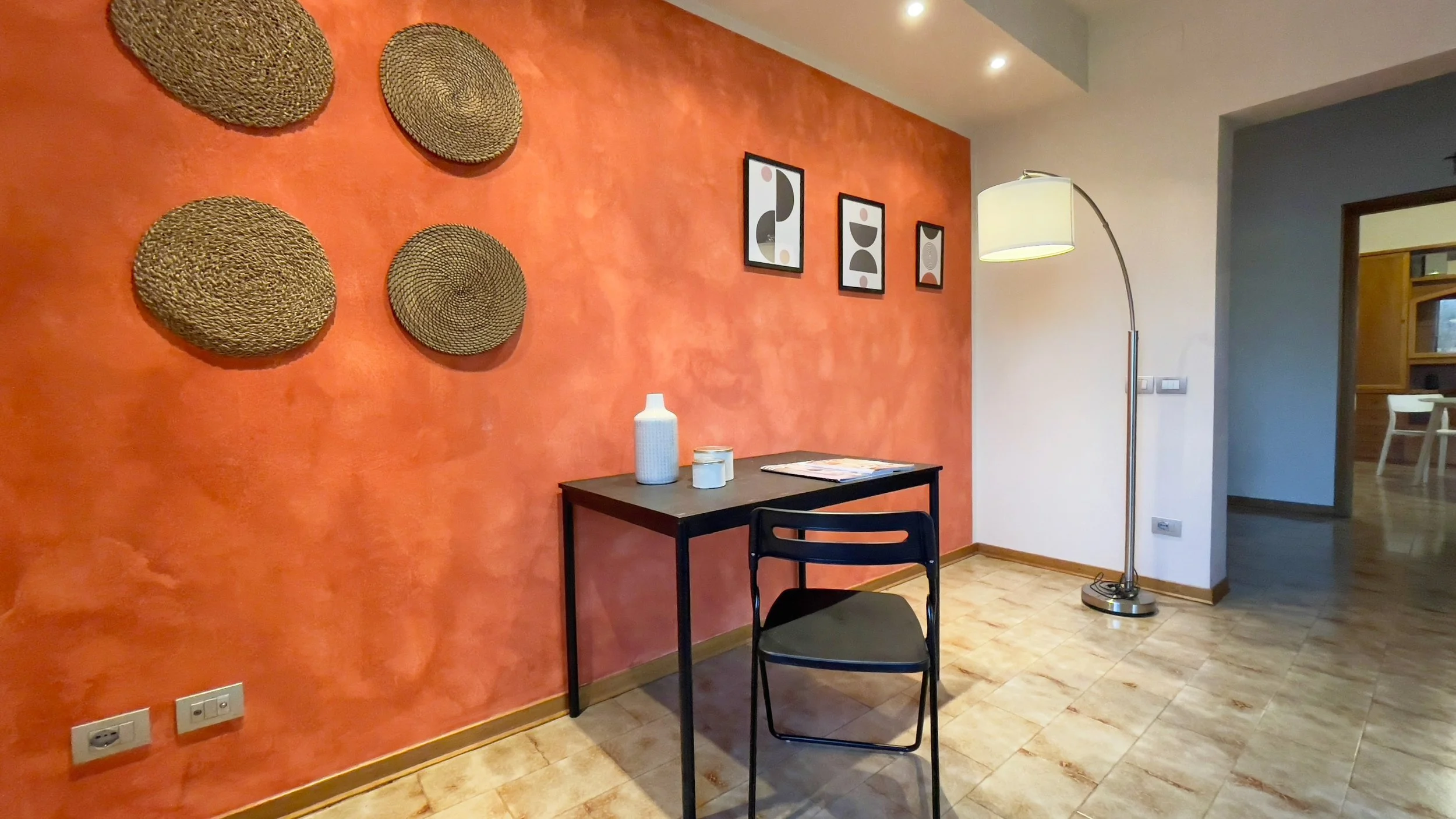 Interno di una stanza con parete arancione decorata con cerchi intrecciati e quadri, un tavolo nero con sedia e lampada da terra.
