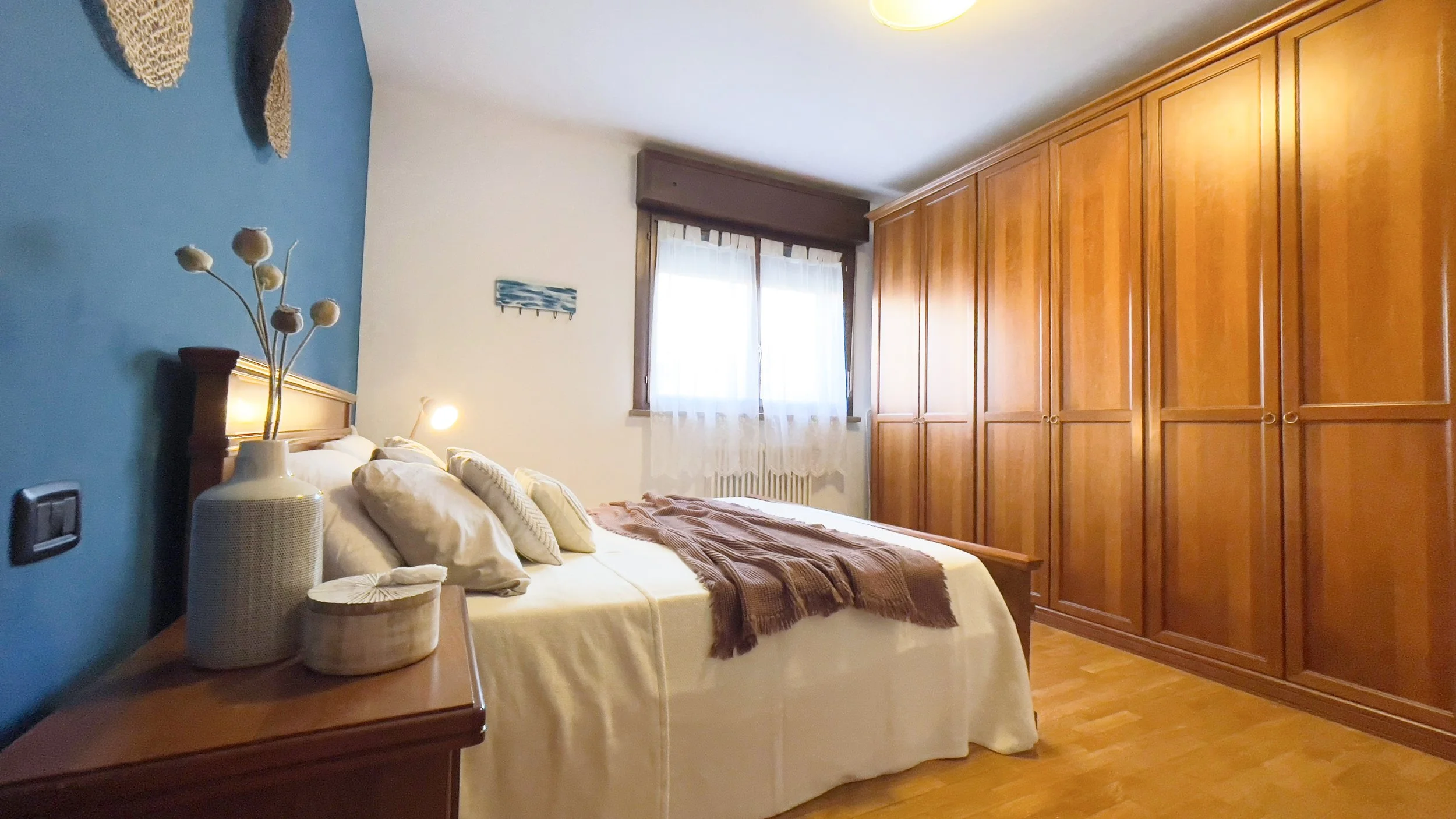 Camera da letto con parete blu, letto matrimoniale con cuscini bianchi e coperta marrone, comodino con vaso e decorazioni, grande armadio in legno, finestra con tendina bianca.