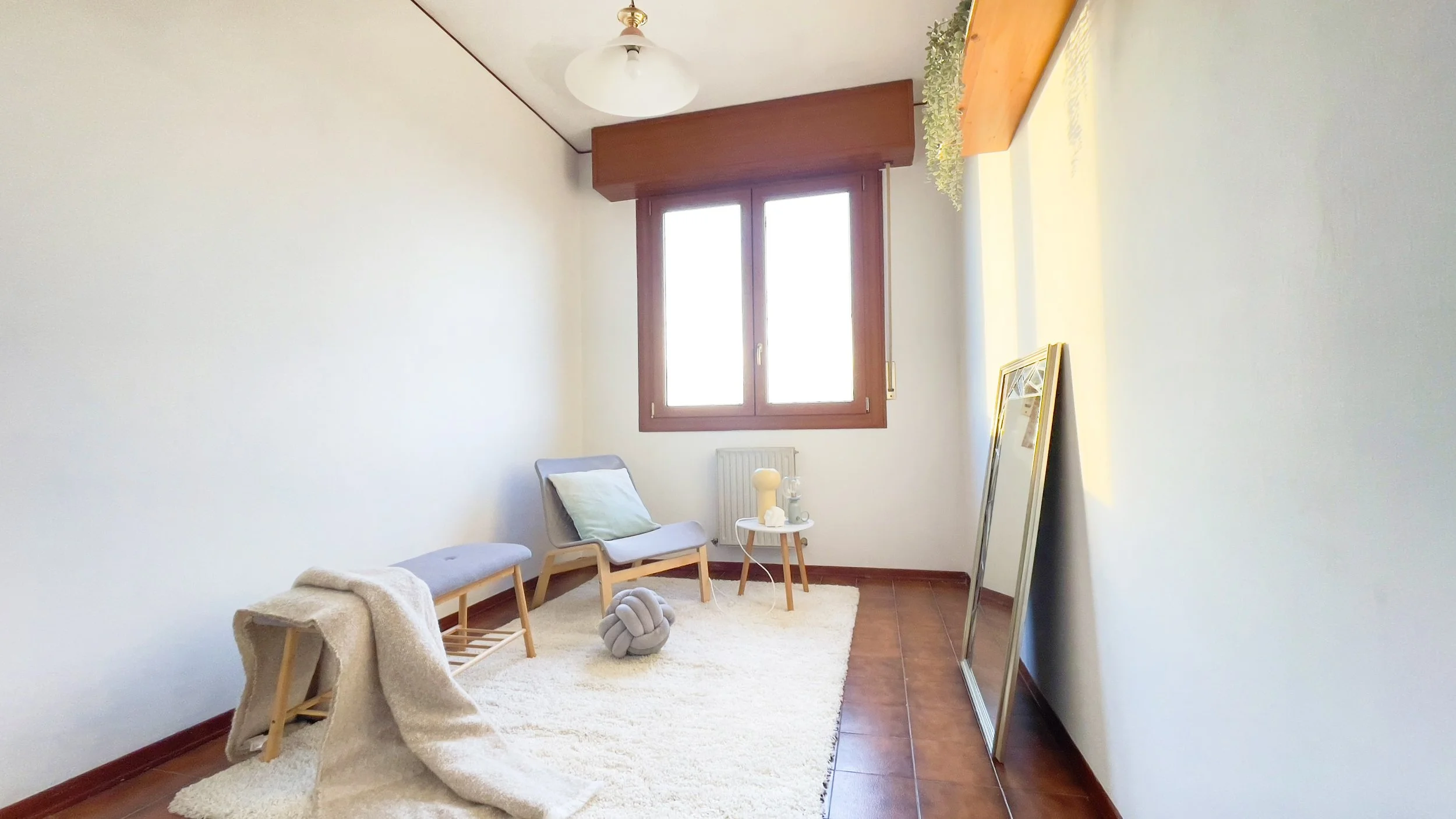 Stanza luminosa con poltrona, tappeto bianco, cuscino, tavolino con lampada, specchio appoggiato e finestra con cornice in legno.