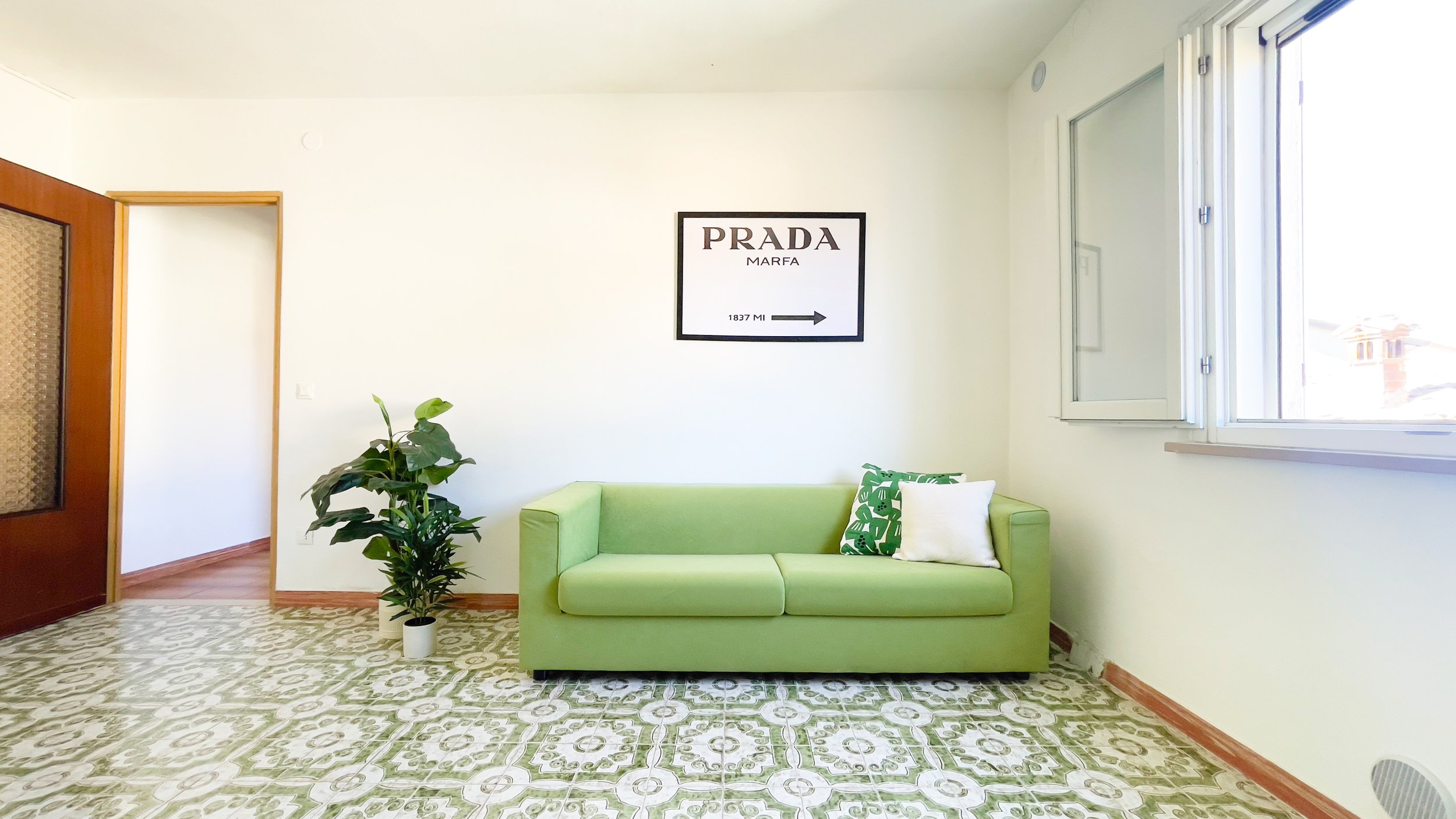 Soggiorno con divano verde, pianta in vaso, finestra e quadro con scritta "PRADA MARFA".